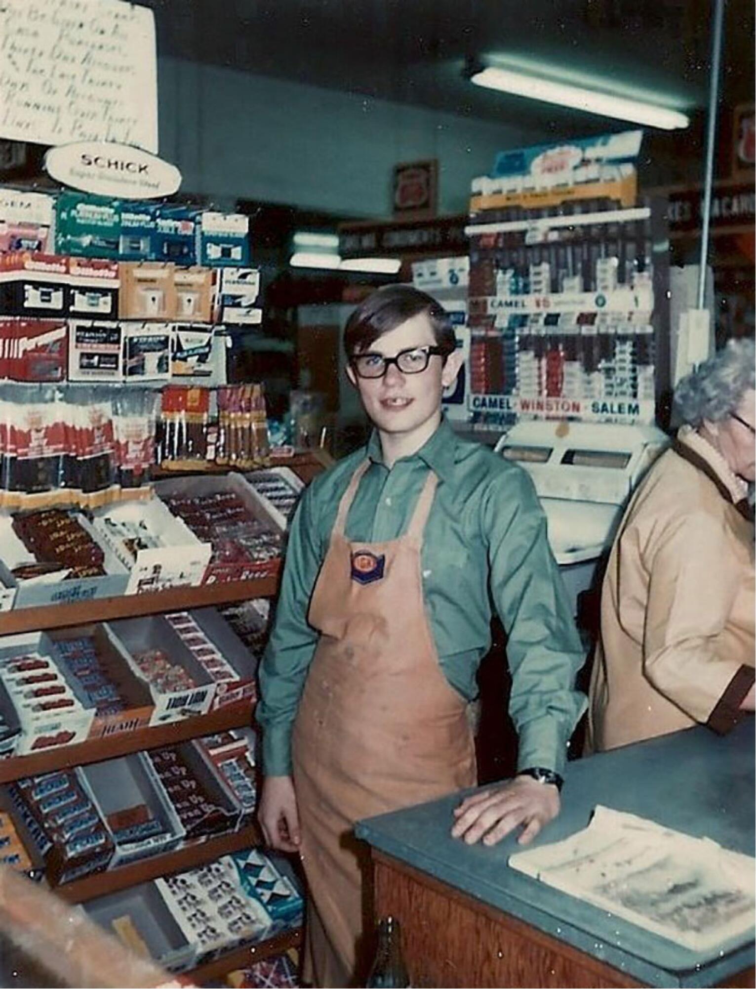 A young man with glasses wearing a green button-down shirt and brown apron stands behind a store counter, with displays of candy and tobacco products visible behind him.