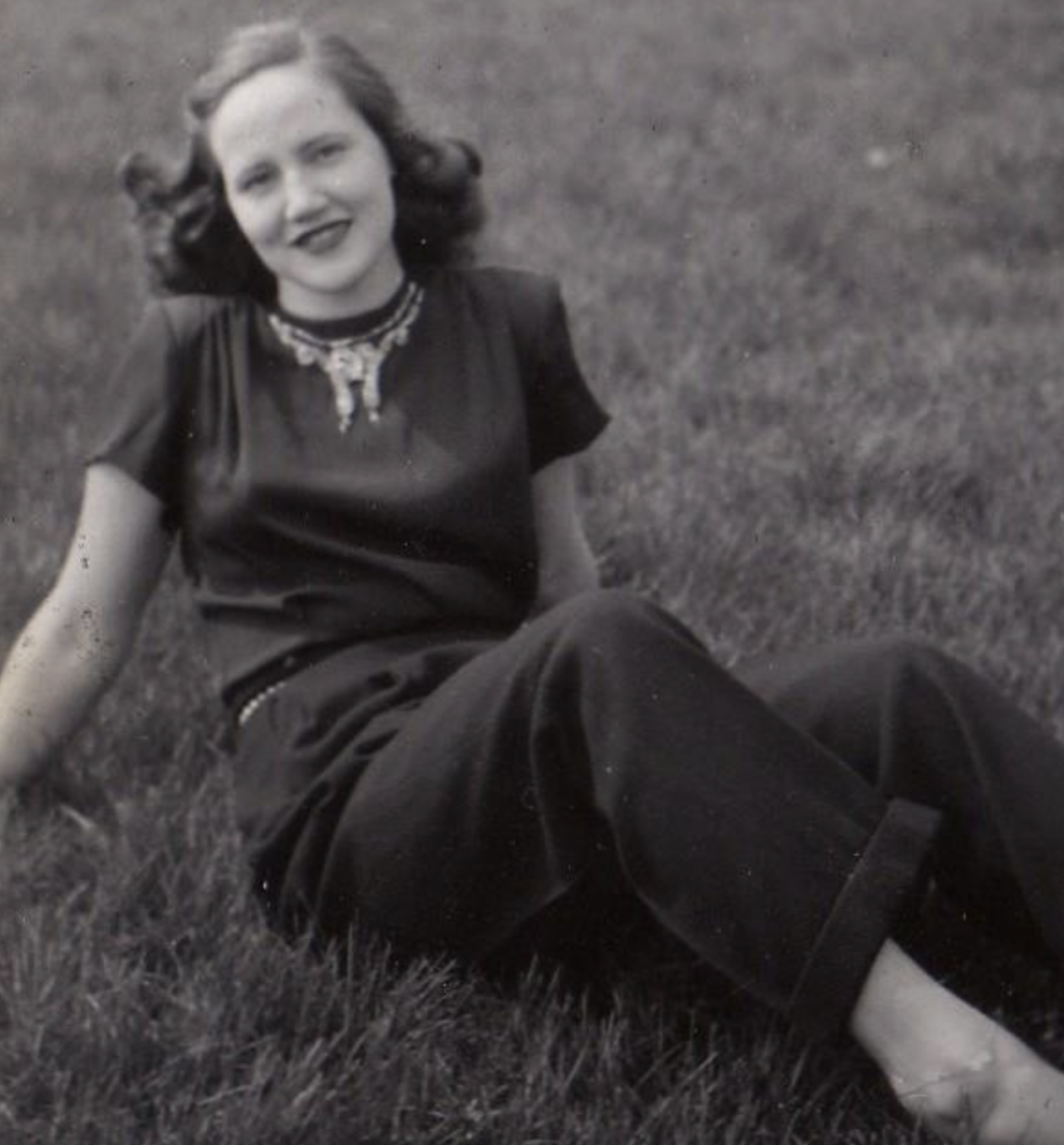 A young woman sitting on grass, wearing a dark short-sleeved top with a decorative necklace.