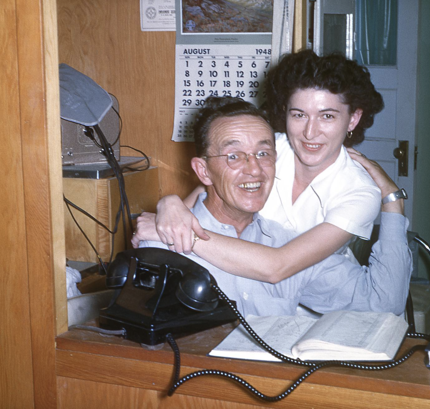 A smiling man and woman pose together at a desk with a rotary telephone and an August 1948 calendar on the wall behind them.