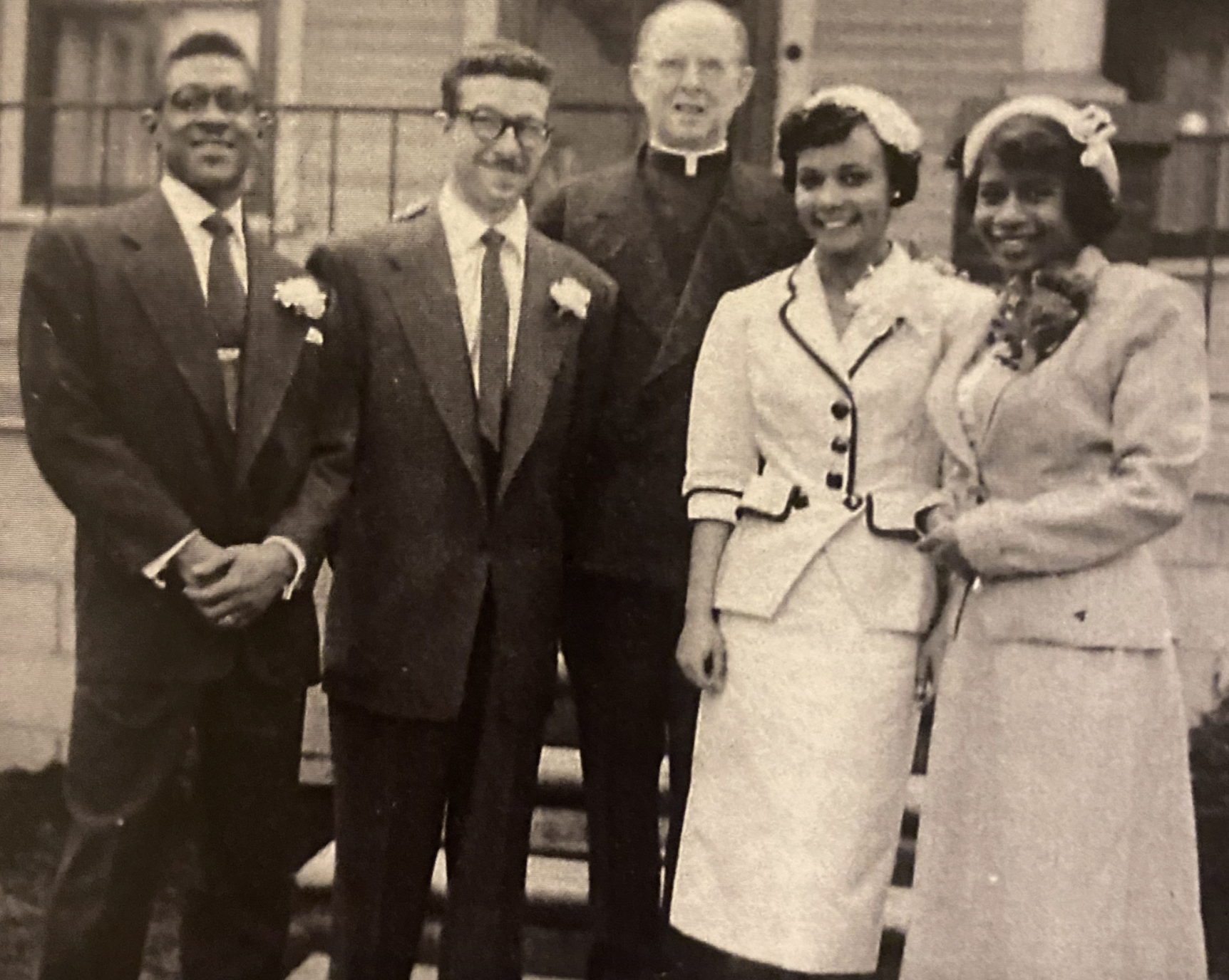 A black-and-white photo of two men in suits, a man in a clerical collar, and two women in formal attire standing together outdoors.
