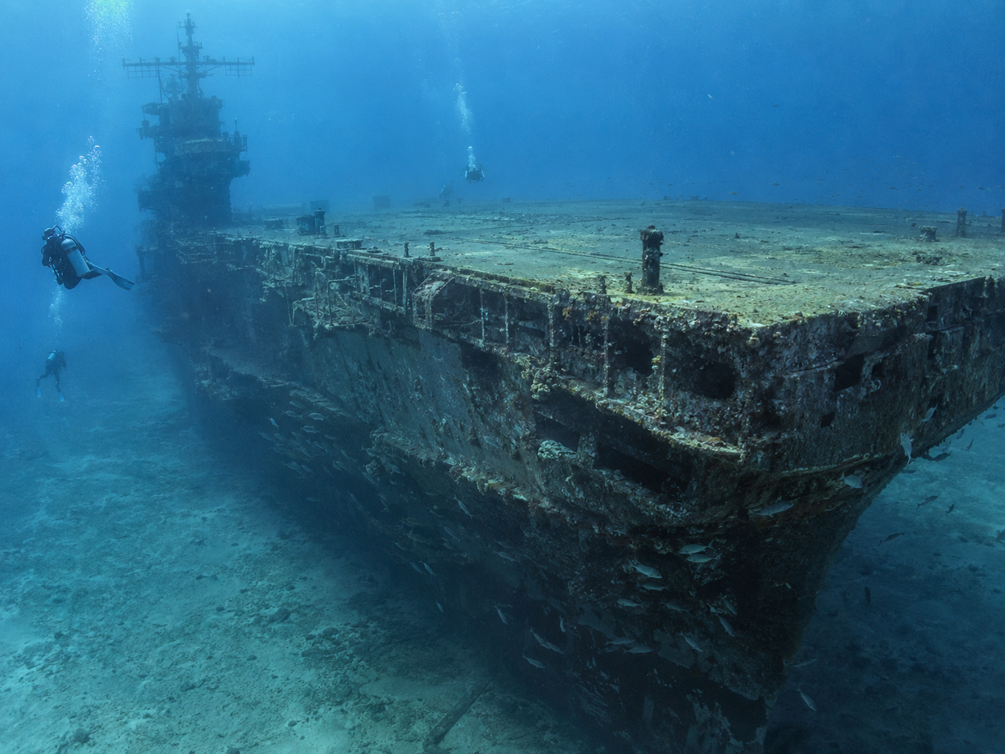 Two scuba divers swim near the massive, sunken remains of an aircraft carrier on the ocean floor.