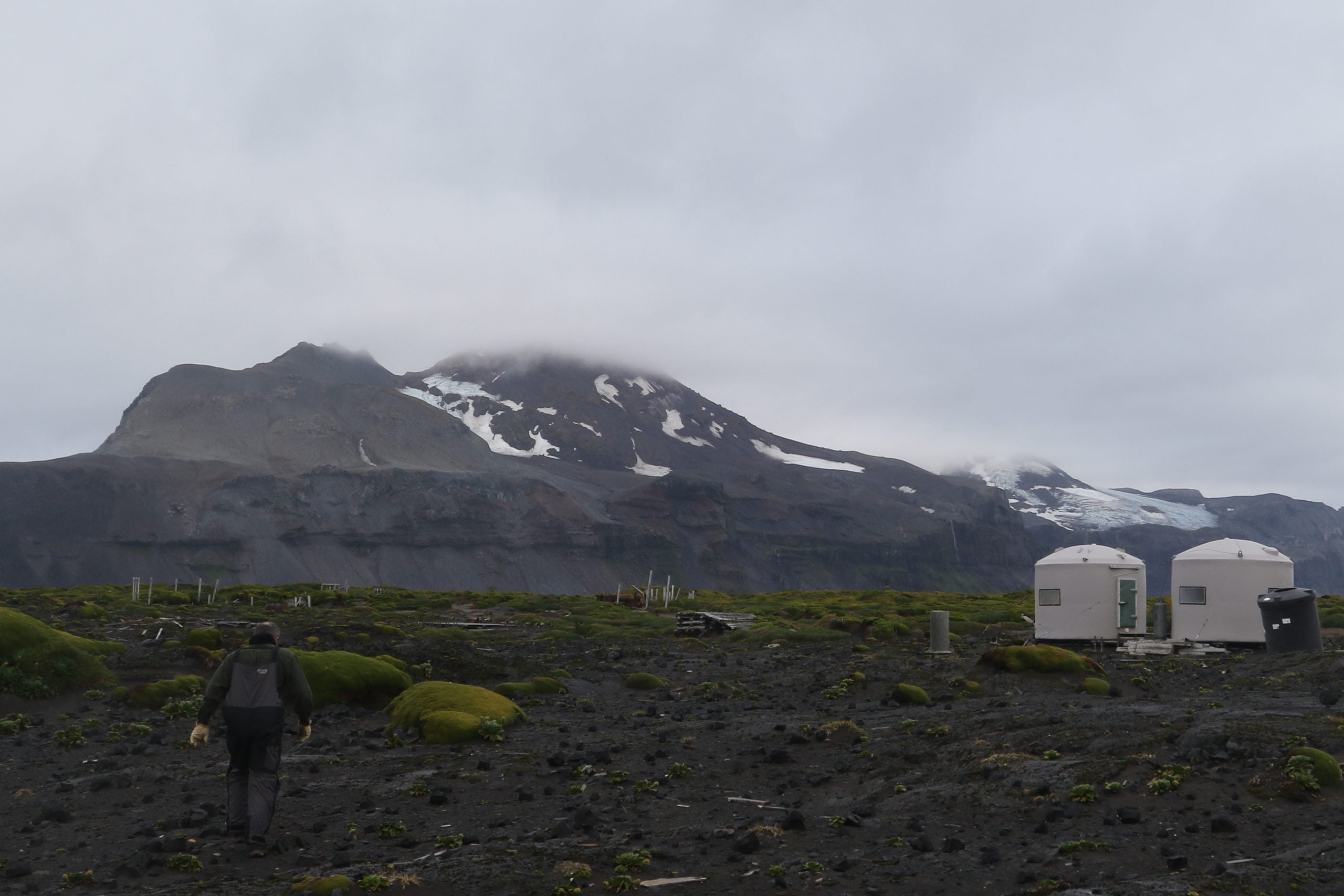A person walks across a rocky, mossy landscape toward two small, white, rounded structures beneath a large, cloudy mountain.