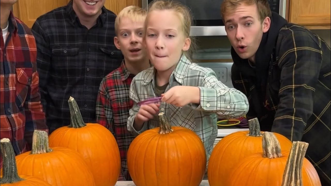 Four people stand behind a counter with several pumpkins, as a young girl in the center prepares to carve one with a small plastic tool.