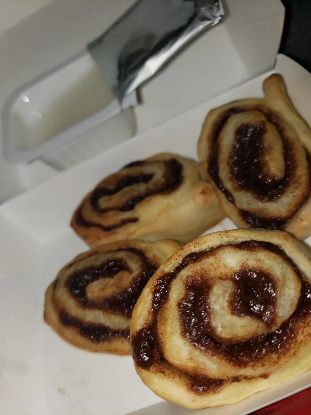 Four cinnamon rolls in a white takeout container next to a small cup of white icing.