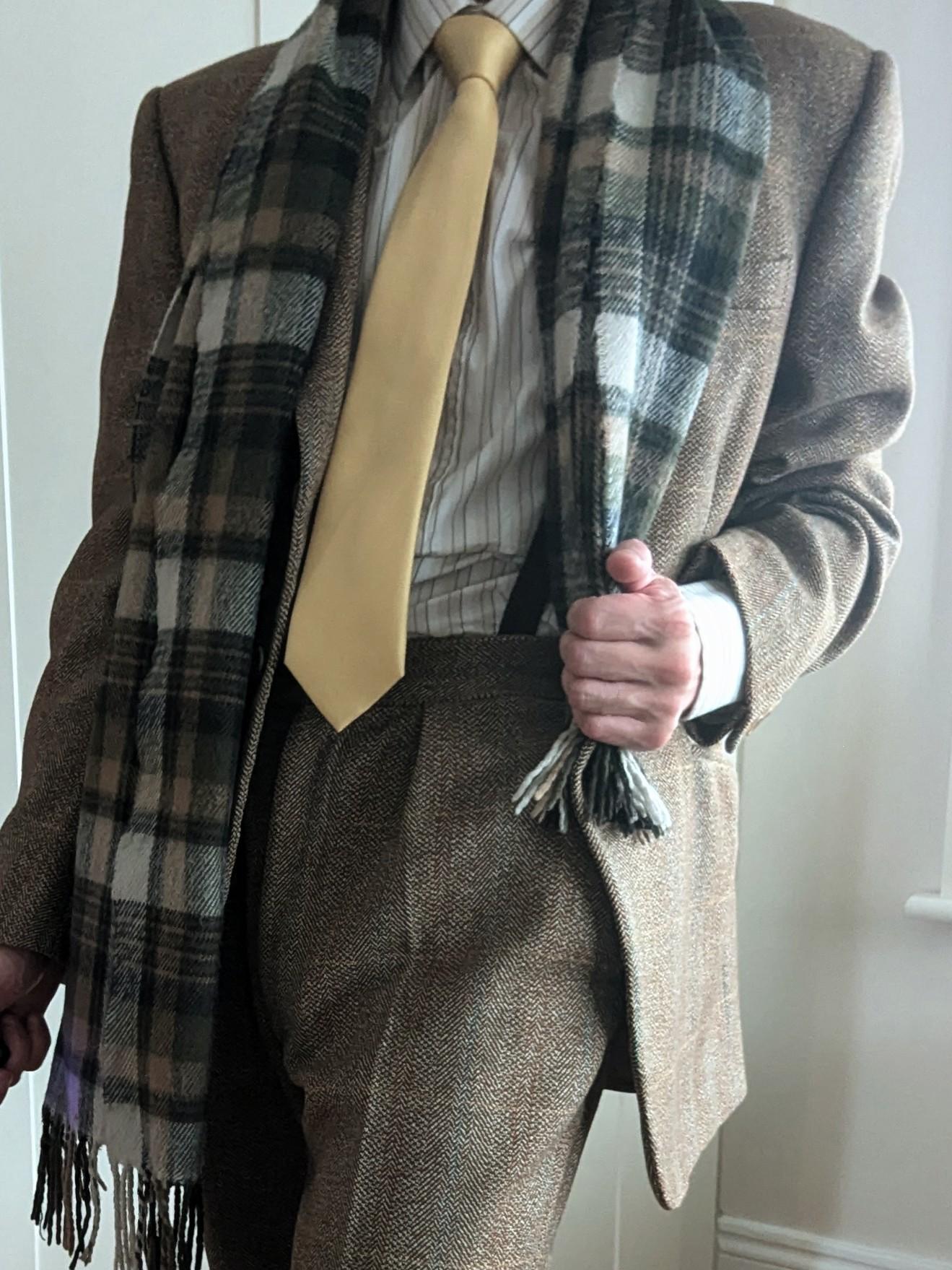 A person wearing a brown suit, yellow tie, striped shirt, and a black-and-white plaid scarf stands indoors, holding one end of the scarf. Their face is out of view.