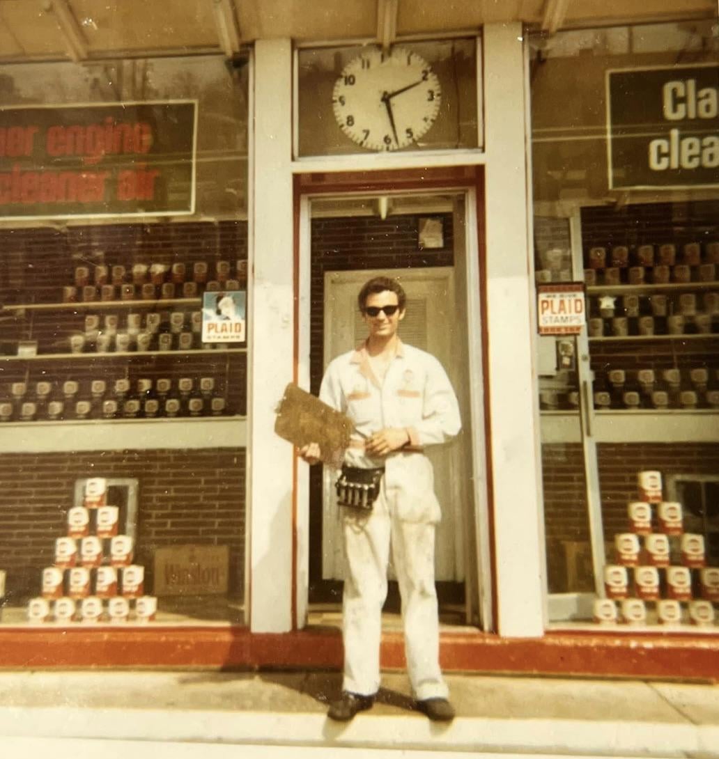 A man in a white uniform and sunglasses stands outside a service station, holding a clipboard in front of a storefront window with stacked oil cans and Plaid Stamps signs.