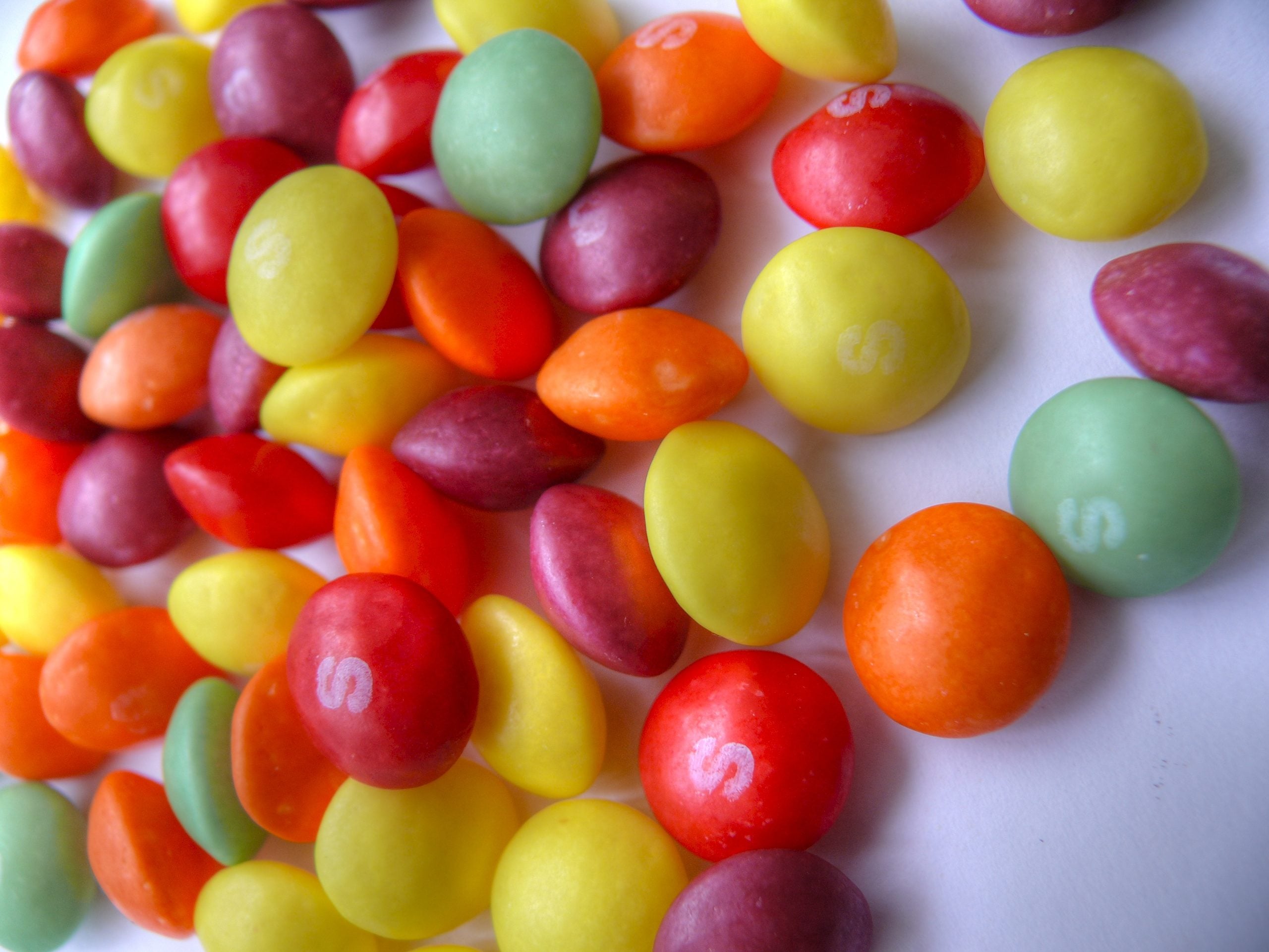 A scattering of multicolored Skittles candies, many marked with the letter “S.”
