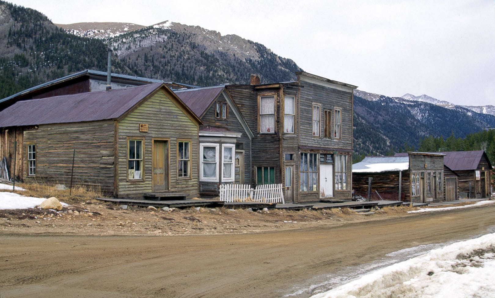 A row of weathered wooden buildings in a historic mountain town with snow-dusted peaks in the background.