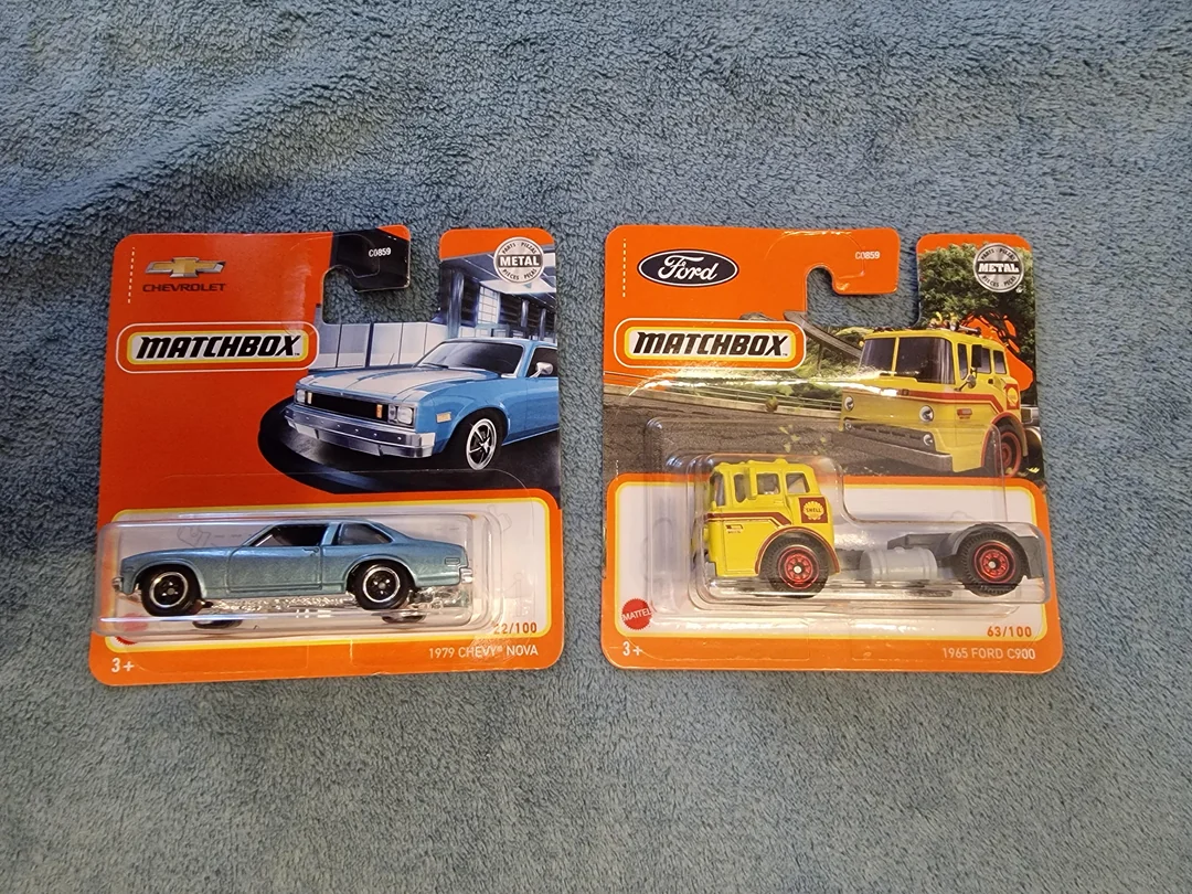 Two Matchbox toy cars in packages on a blue towel: a blue 1979 Chevy Nova on the left and a yellow 1965 Ford C900 tow truck on the right. Both packages have orange backgrounds and brand logos.