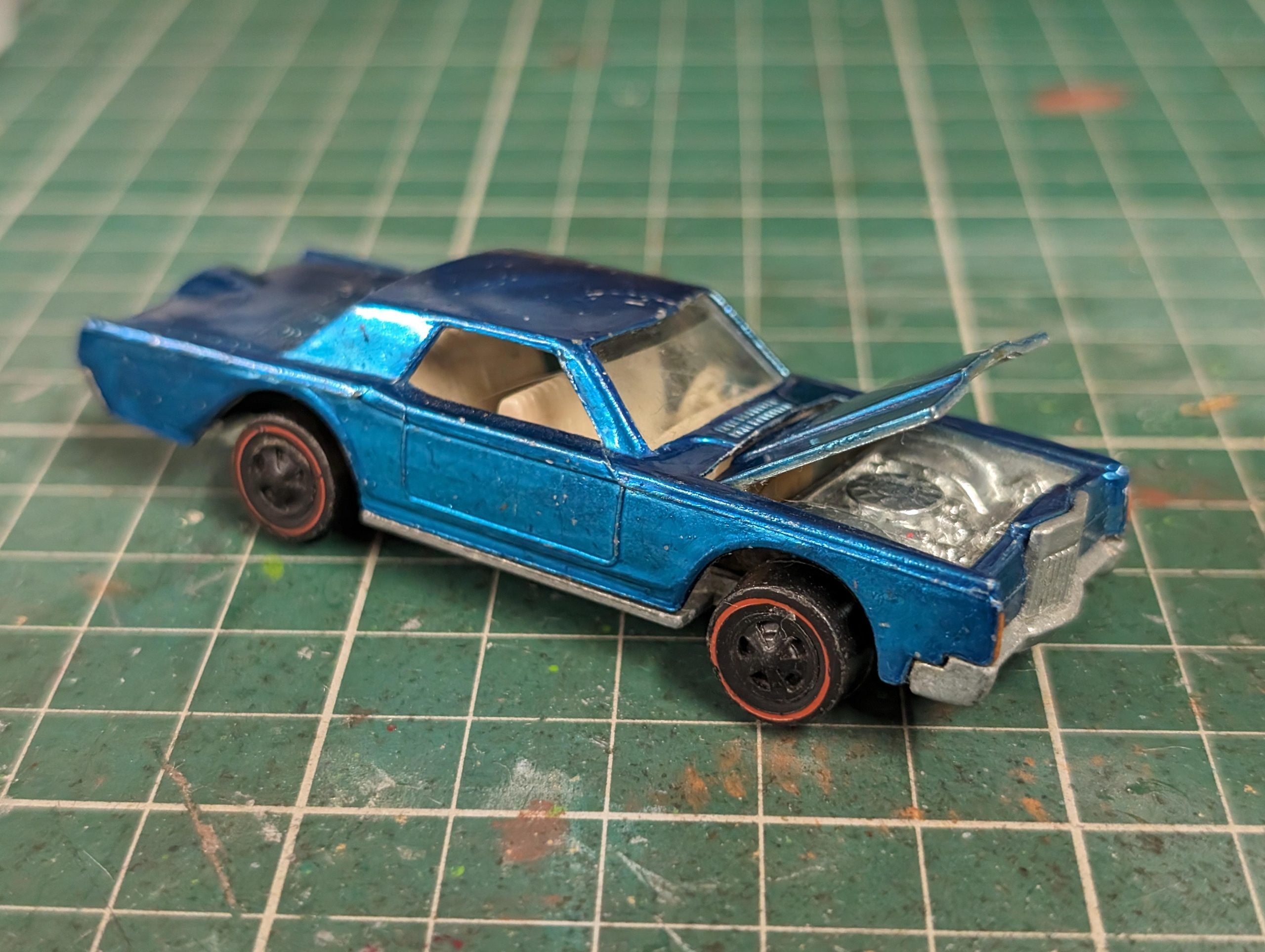 A blue toy car with an open hood sits on a green cutting mat. The car has worn paint, red-lined wheels, and visible engine details.