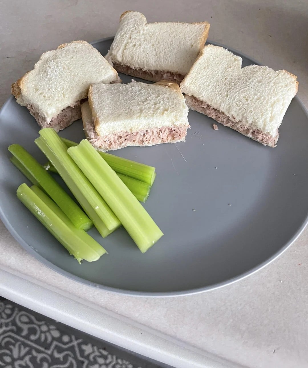 A gray plate with four small tuna sandwiches on white bread and several celery sticks, placed on a light-colored countertop.