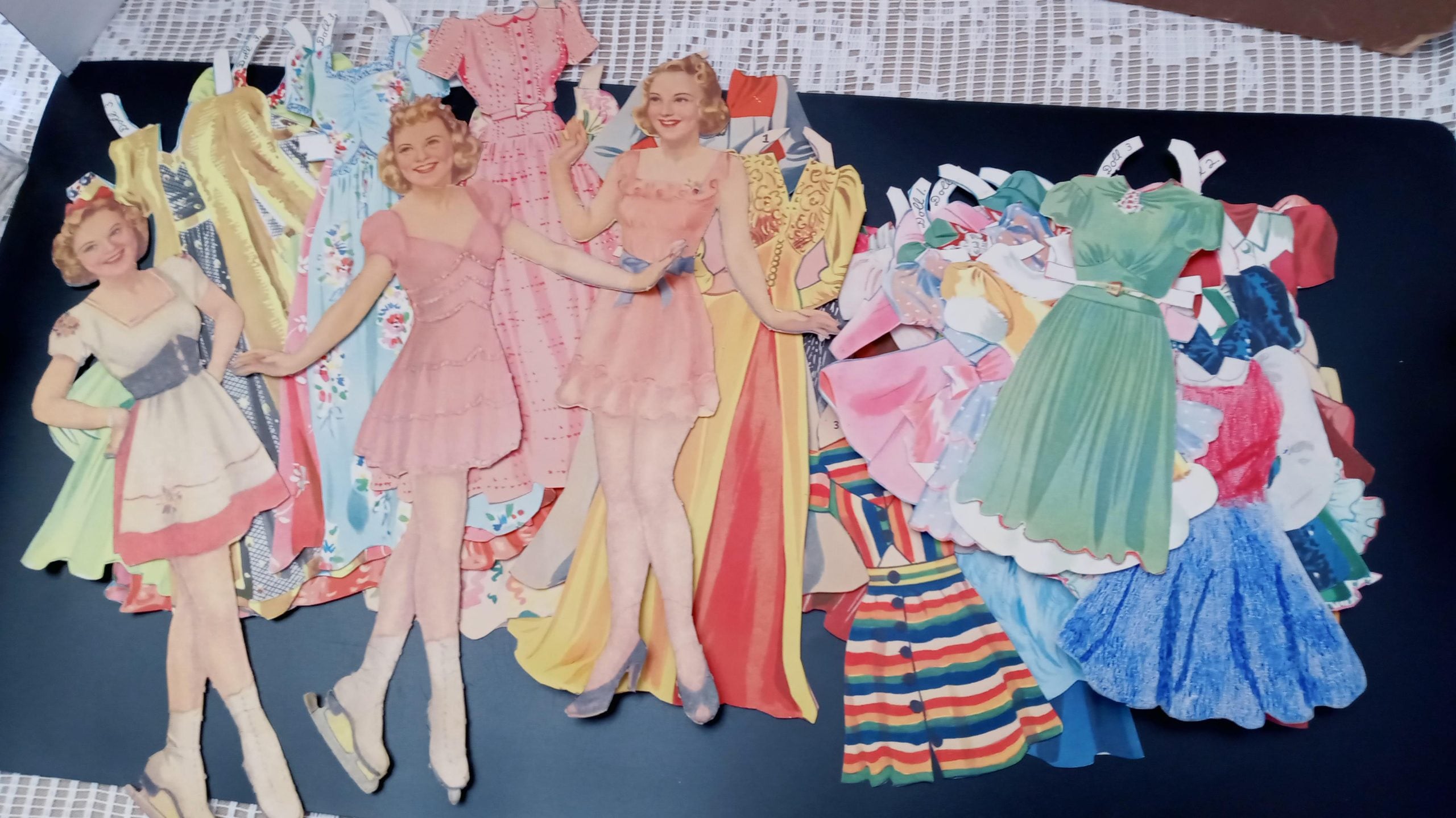 Two vintage paper dolls with an assortment of illustrated paper dresses and outfits spread out on a dark surface.
