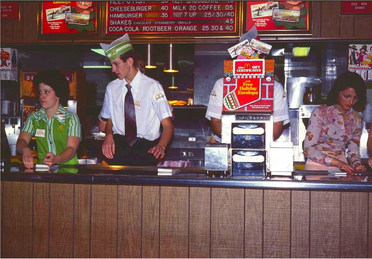 Three McDonald’s employees work behind a counter beneath a menu board listing 1970s-era prices for burgers, shakes, and coffee.