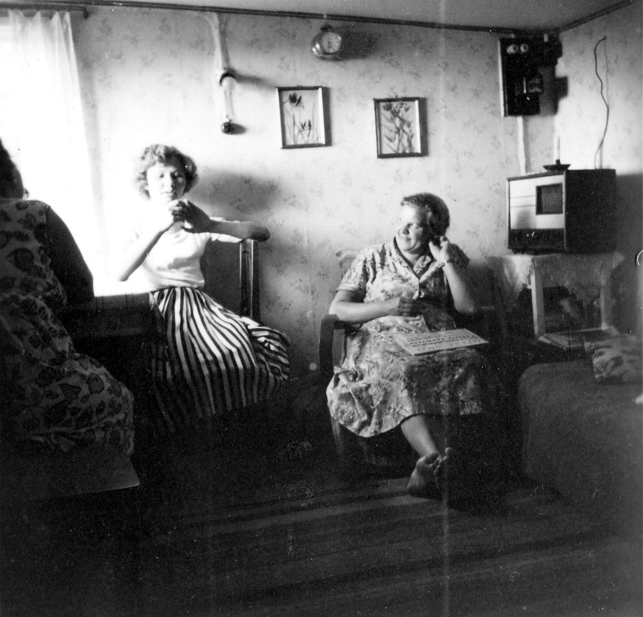 Three women sitting in a living room, with one woman in a striped skirt holding a cup and another woman in a patterned dress sitting in an armchair.