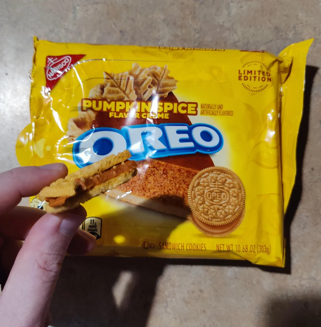 A hand holds a broken Pumpkin Spice Oreo cookie in front of its yellow limited edition packaging.