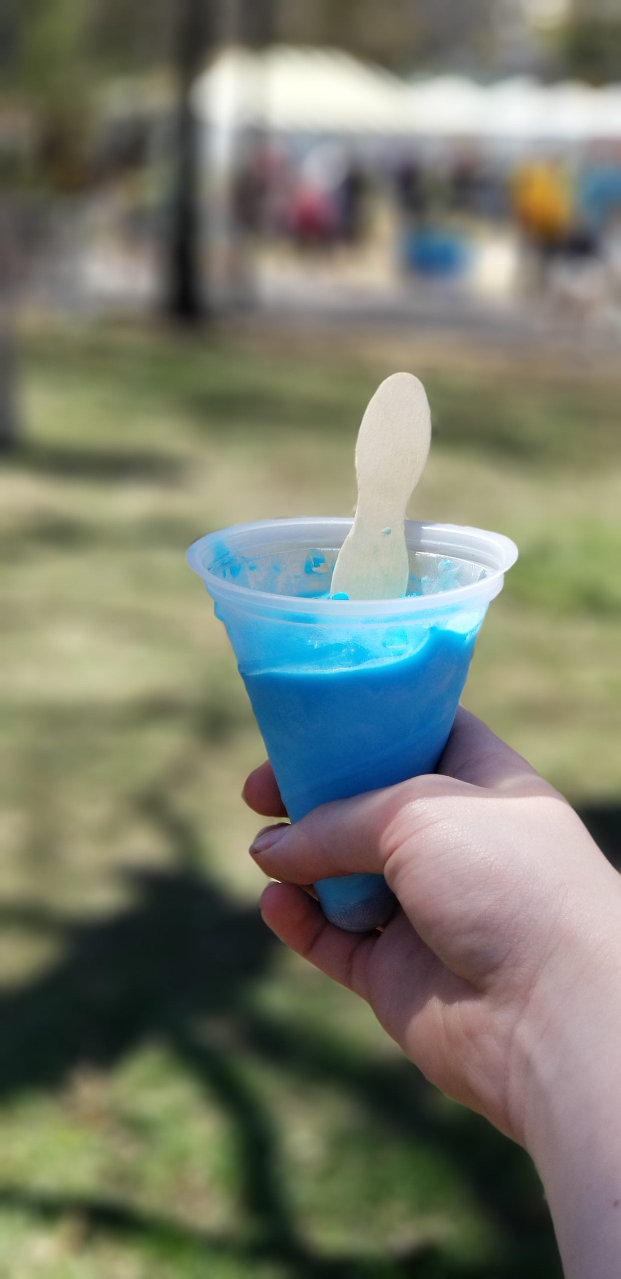 A hand holding a clear plastic cup filled with bright blue shaved ice and a wooden spoon.