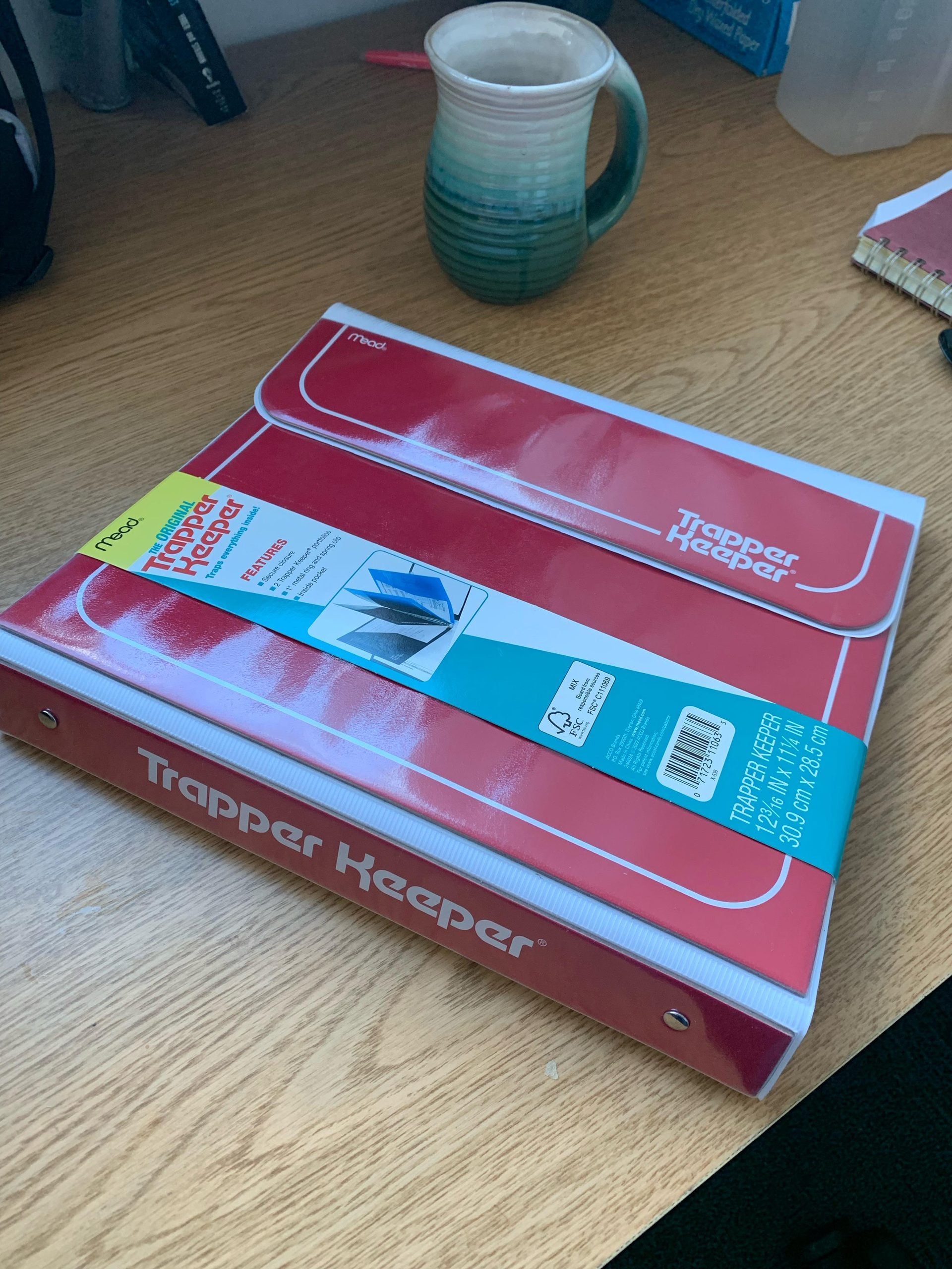 A red Mead Trapper Keeper binder with a product information sleeve lying on a wooden desk next to a teal mug.