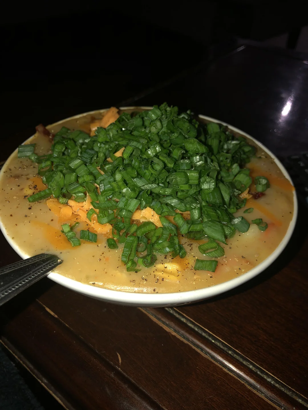A bowl of creamy soup or stew topped with chopped green onions, shredded cheese, and black pepper, set on a dark wooden table.