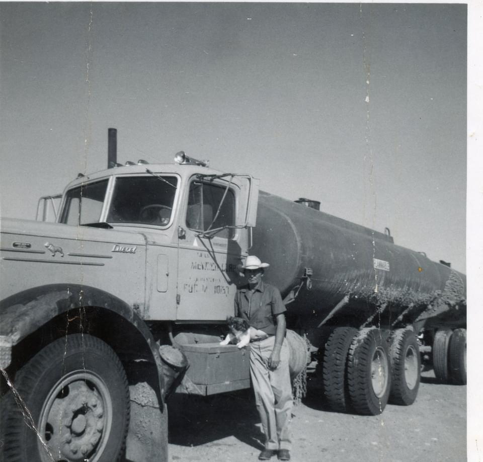 A man wearing a cowboy hat stands next to a tanker truck with a small dog in his arms.