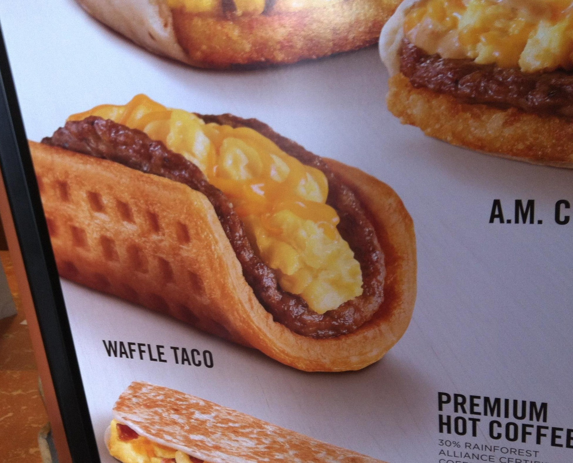 A waffle taco filled with a sausage patty and scrambled eggs, displayed on a menu board.