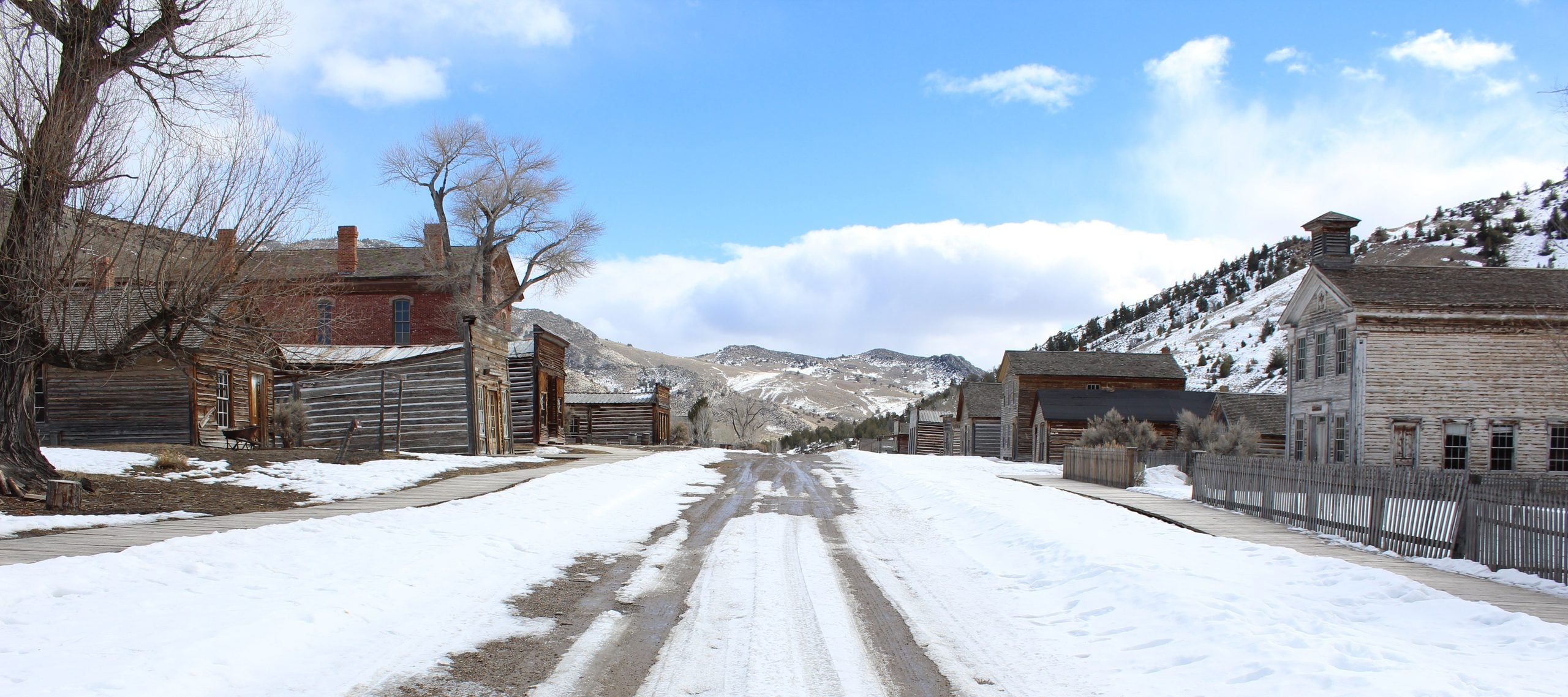 A snow-covered dirt road winds through a historic ghost town with weathered wooden buildings under a bright blue sky.
