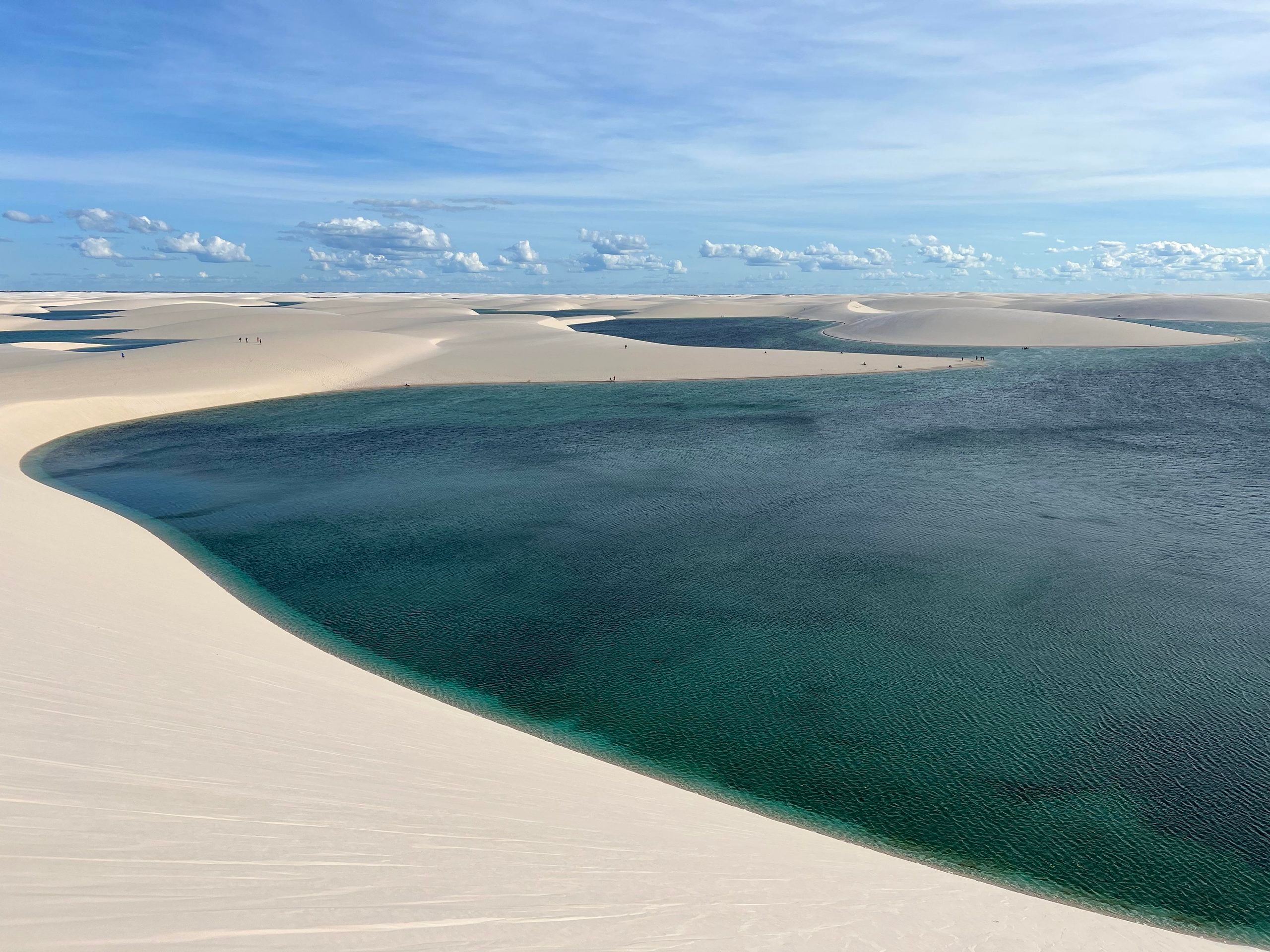 A large, deep blue lagoon surrounded by vast white sand dunes under a blue sky with scattered clouds.
