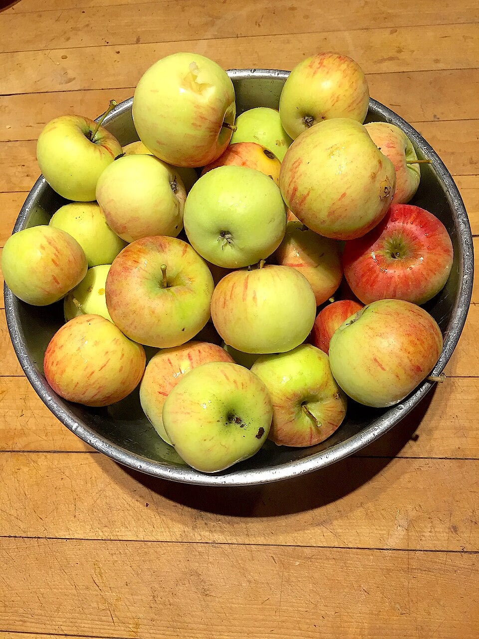 A metal bowl filled with a variety of yellow, green, and red apples sitting on a wooden surface.
