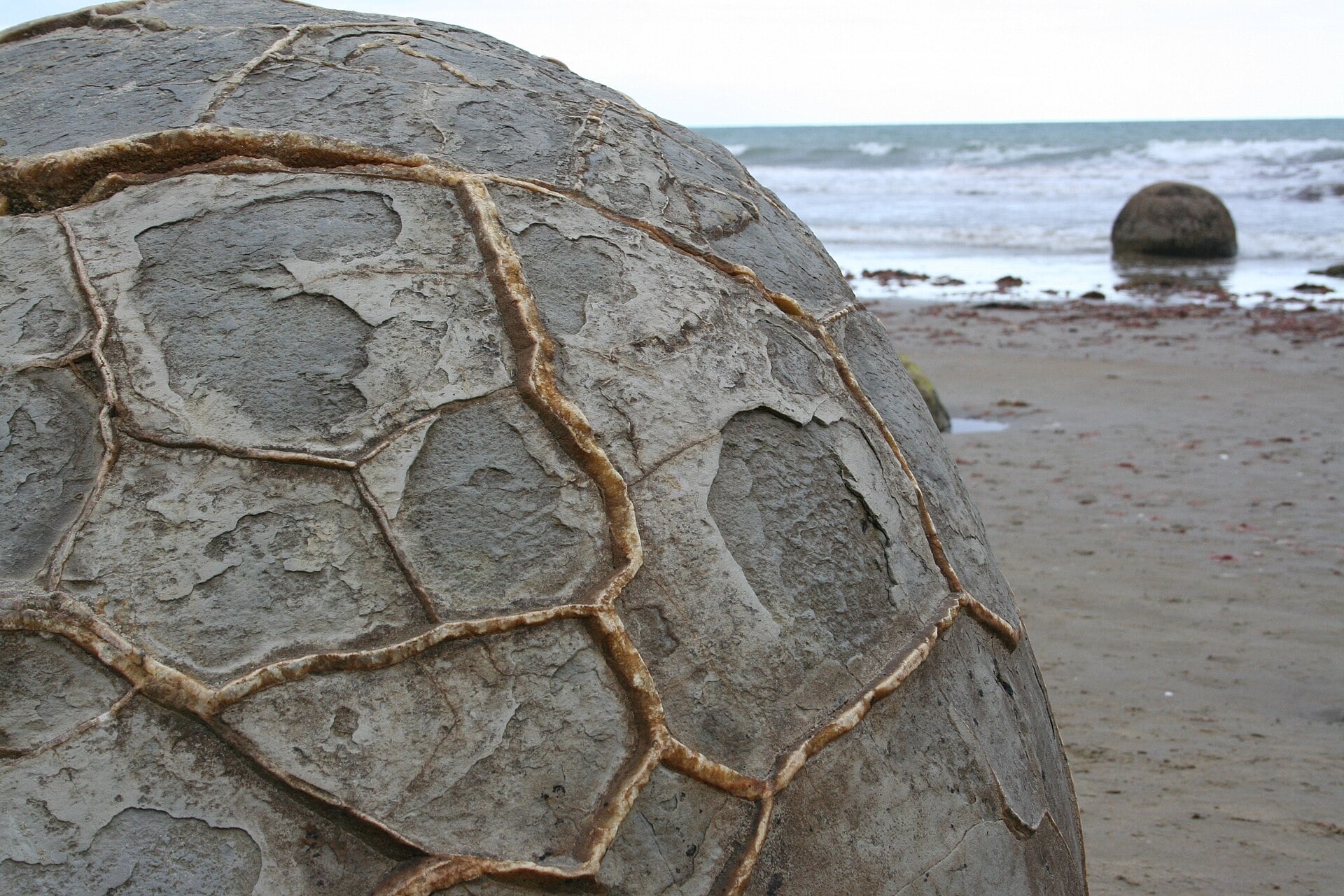 Large, spherical Moeraki boulder with a cracked, textured surface on a sandy beach near the ocean.