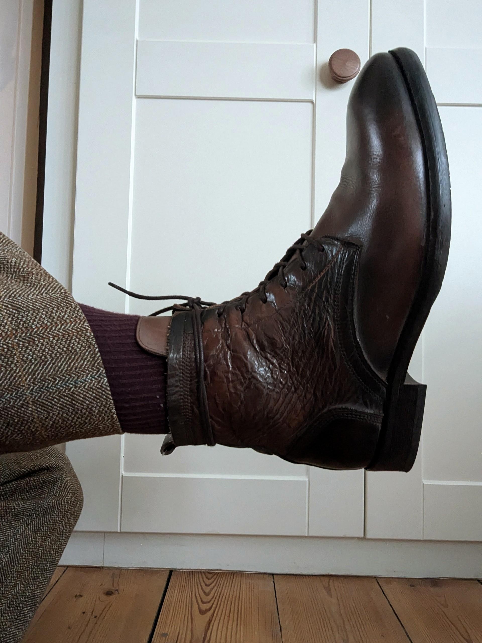 A person wears brown leather lace-up boots, purple socks, and brown patterned pants, with their leg raised in front of white cabinet doors and a wooden floor.