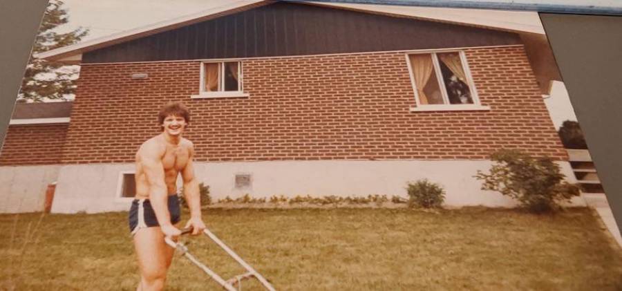 A shirtless man in athletic shorts smiles while pushing a lawnmower in front of a brick house.