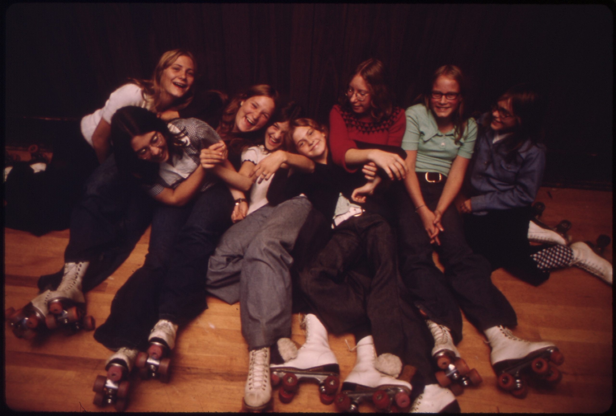 Seven teenage girls in casual clothes sitting together on a wooden roller rink floor, all wearing roller skates.
