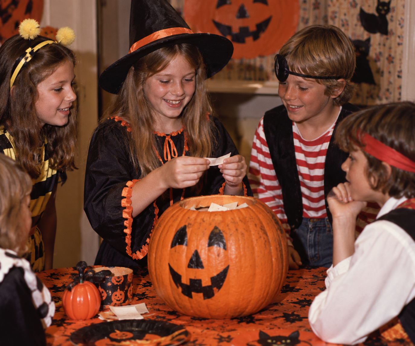 Four children in Halloween costumes gather around a carved pumpkin on a table, watching as a girl in a witch hat reads a slip of paper from inside it.