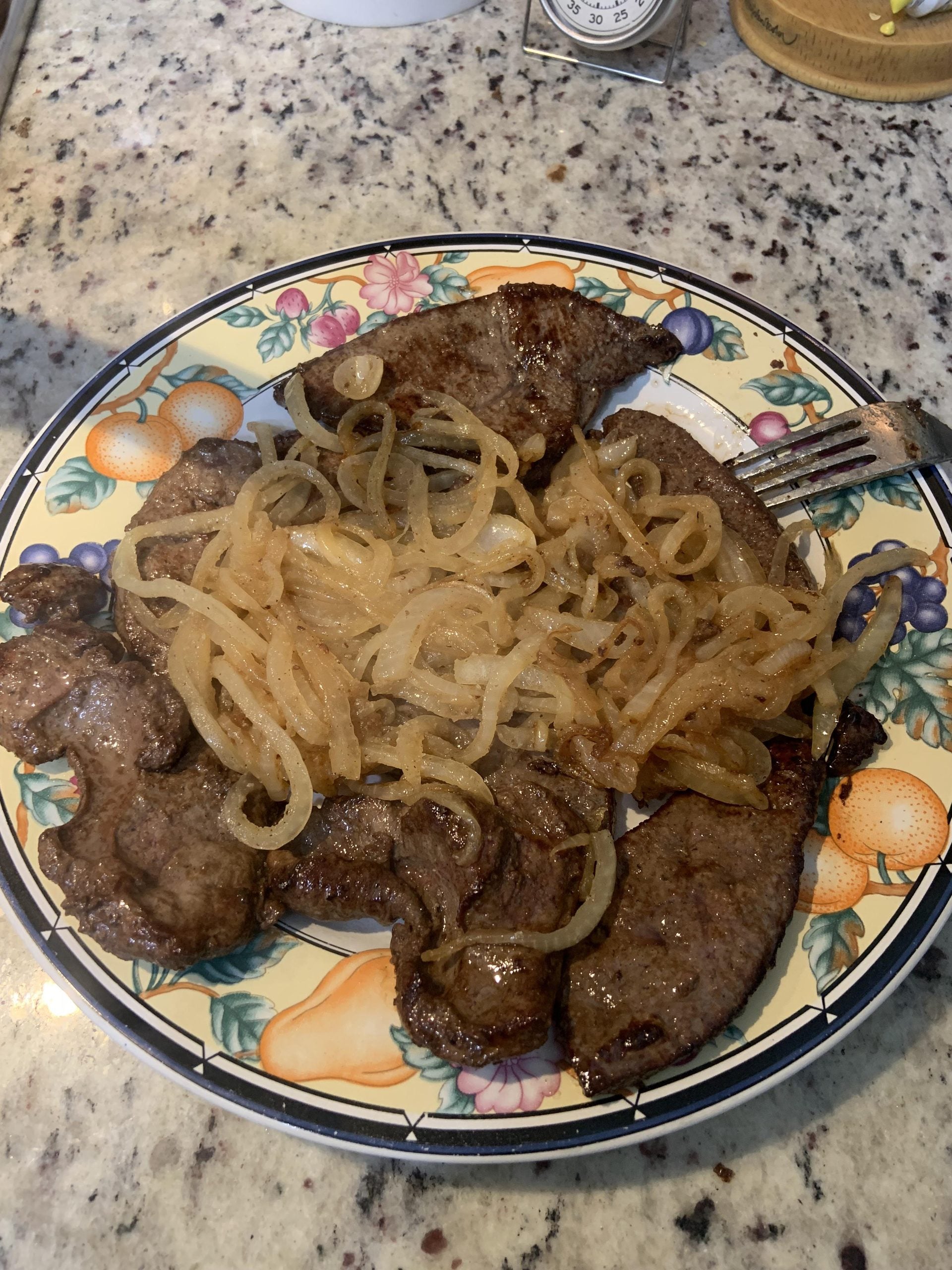 A plate with cooked liver pieces topped with sautéed onions sits on a floral-patterned dish atop a granite countertop.