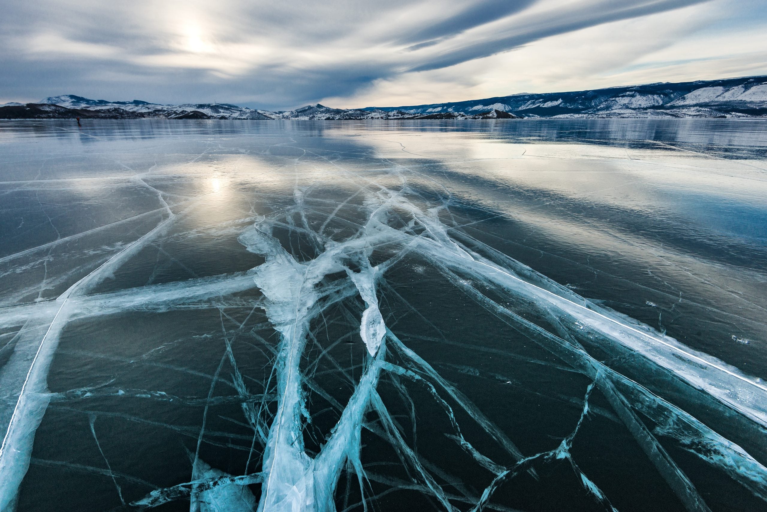 Wide expanse of ice on Lake Baikal featuring intricate, deep cracks, with snow-covered mountains in the distance under a cloudy sky.