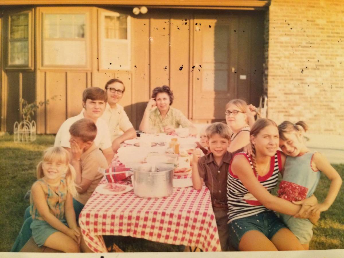 A family sits around a picnic table with a red-and-white checkered cloth in a backyard.