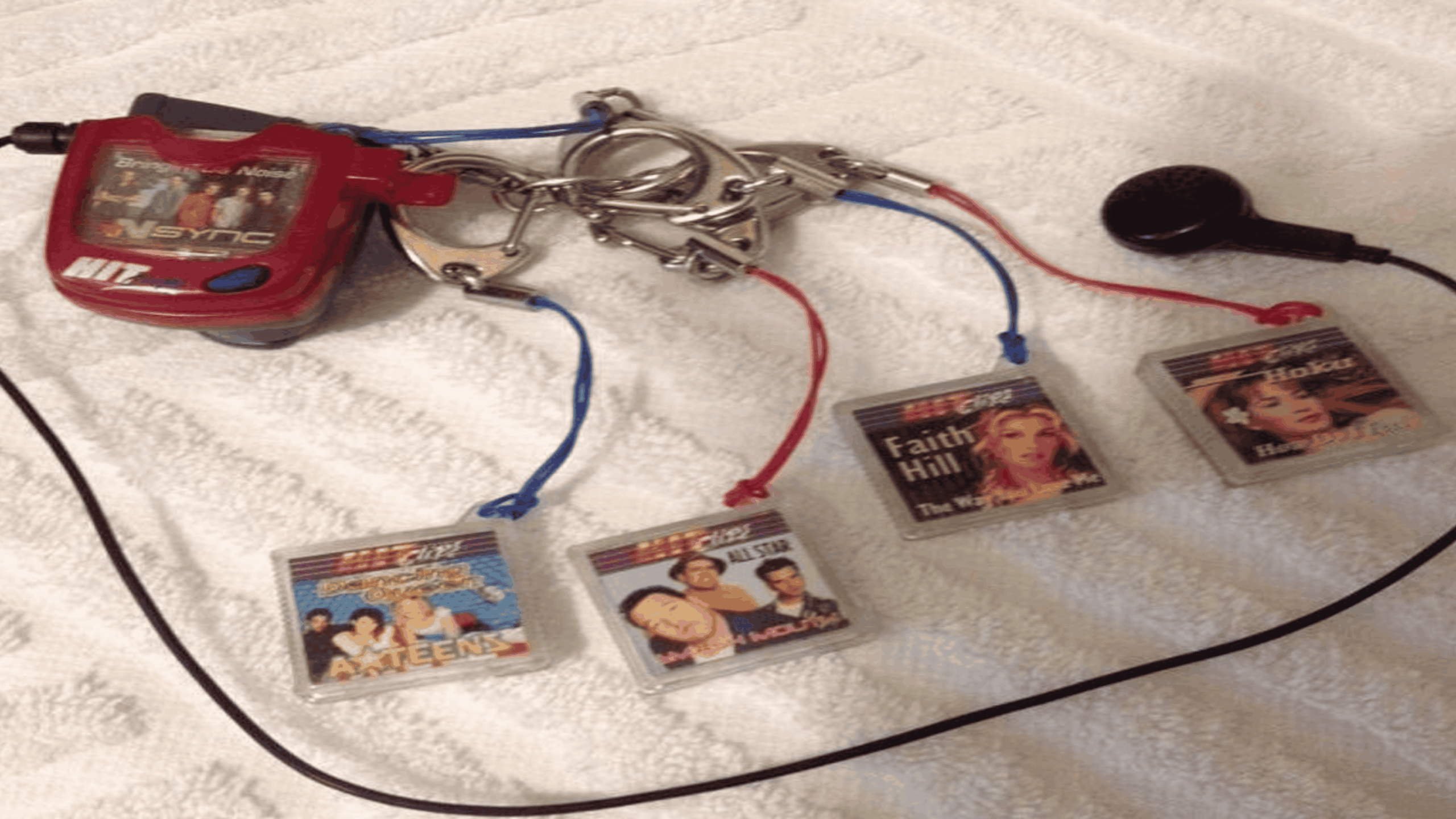 A red HitClips player with four attached music cartridges featuring artwork for NSYNC, A-Teens, Smash Mouth, Faith Hill, and Hoku, plus an attached earbud.