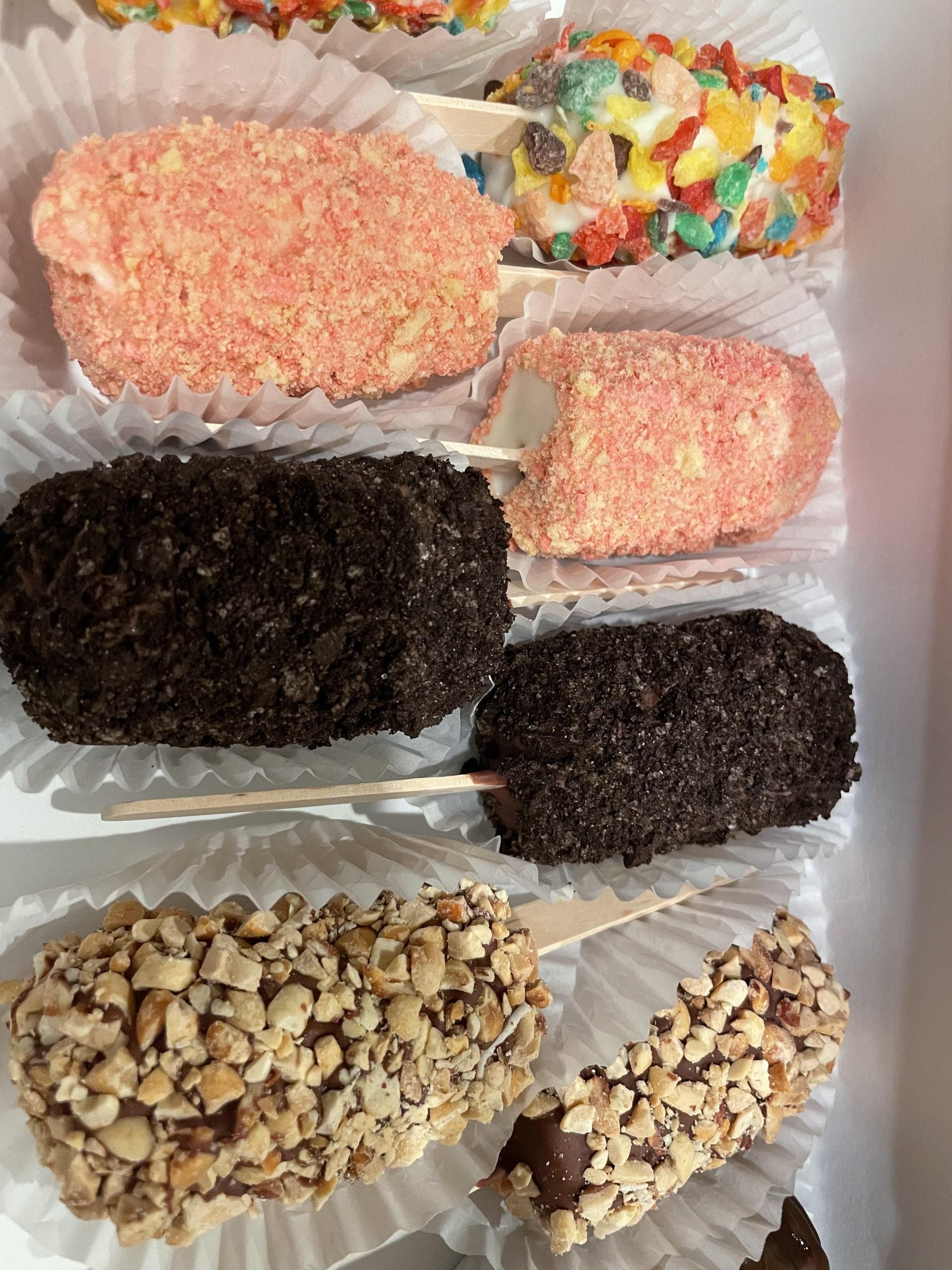 A box of ice cream bars on sticks, coated in varied toppings including crushed cookies, nuts, pink cake crumbs, and fruity cereal.