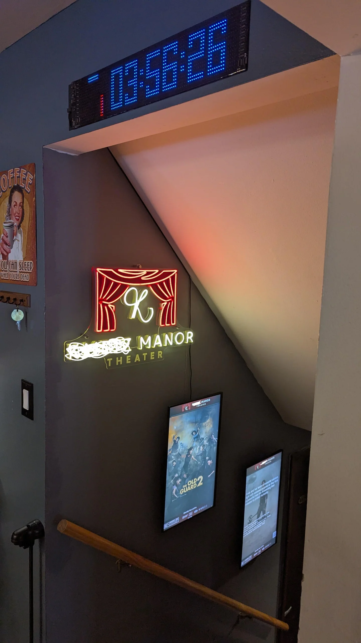 A staircase wall featuring a digital clock, a “K Manor Theater” neon sign, and two movie posters, one for “The Old Guard 2.”