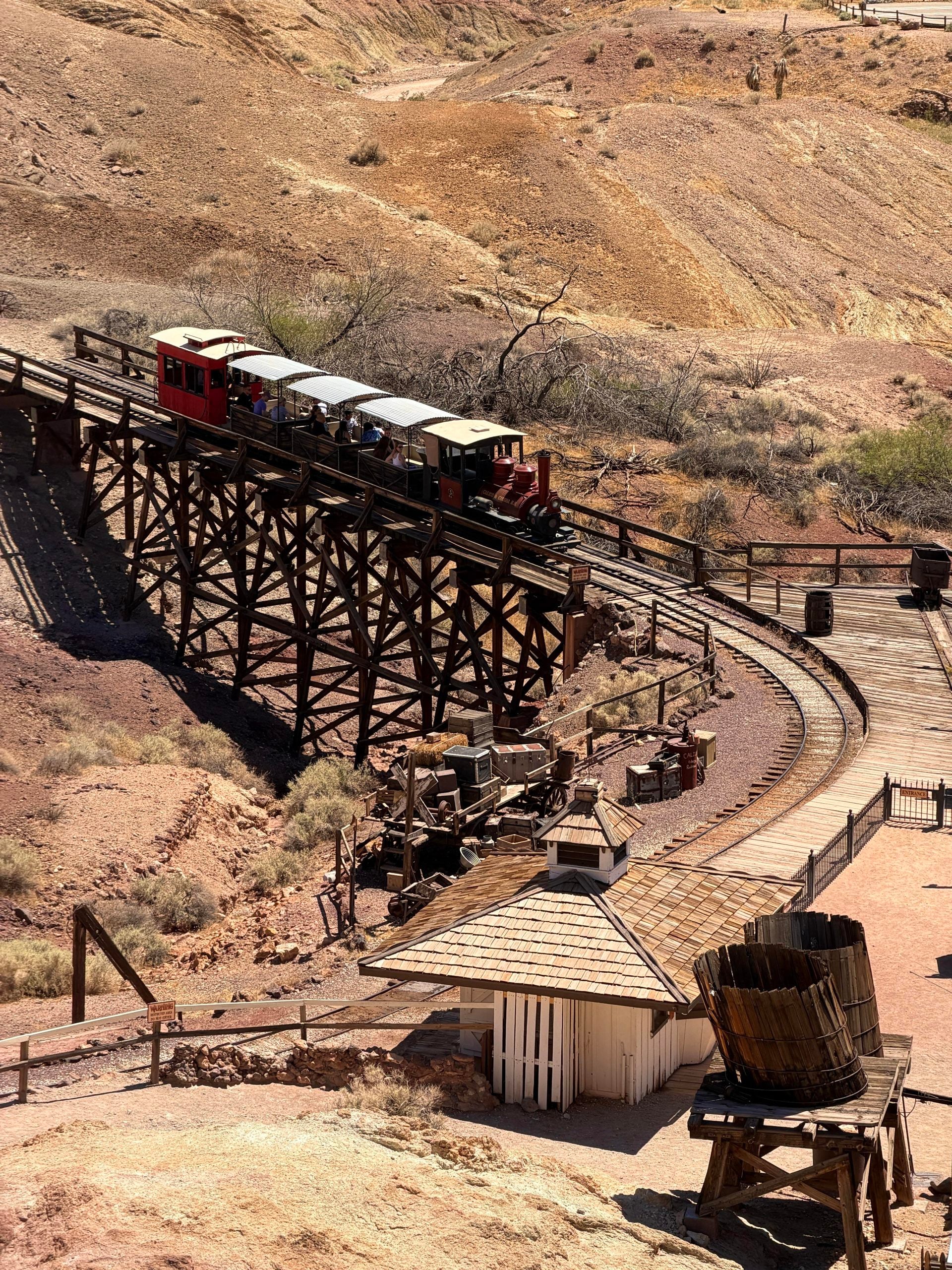 A small red passenger train travels along a wooden trestle bridge through a dry, hilly landscape.