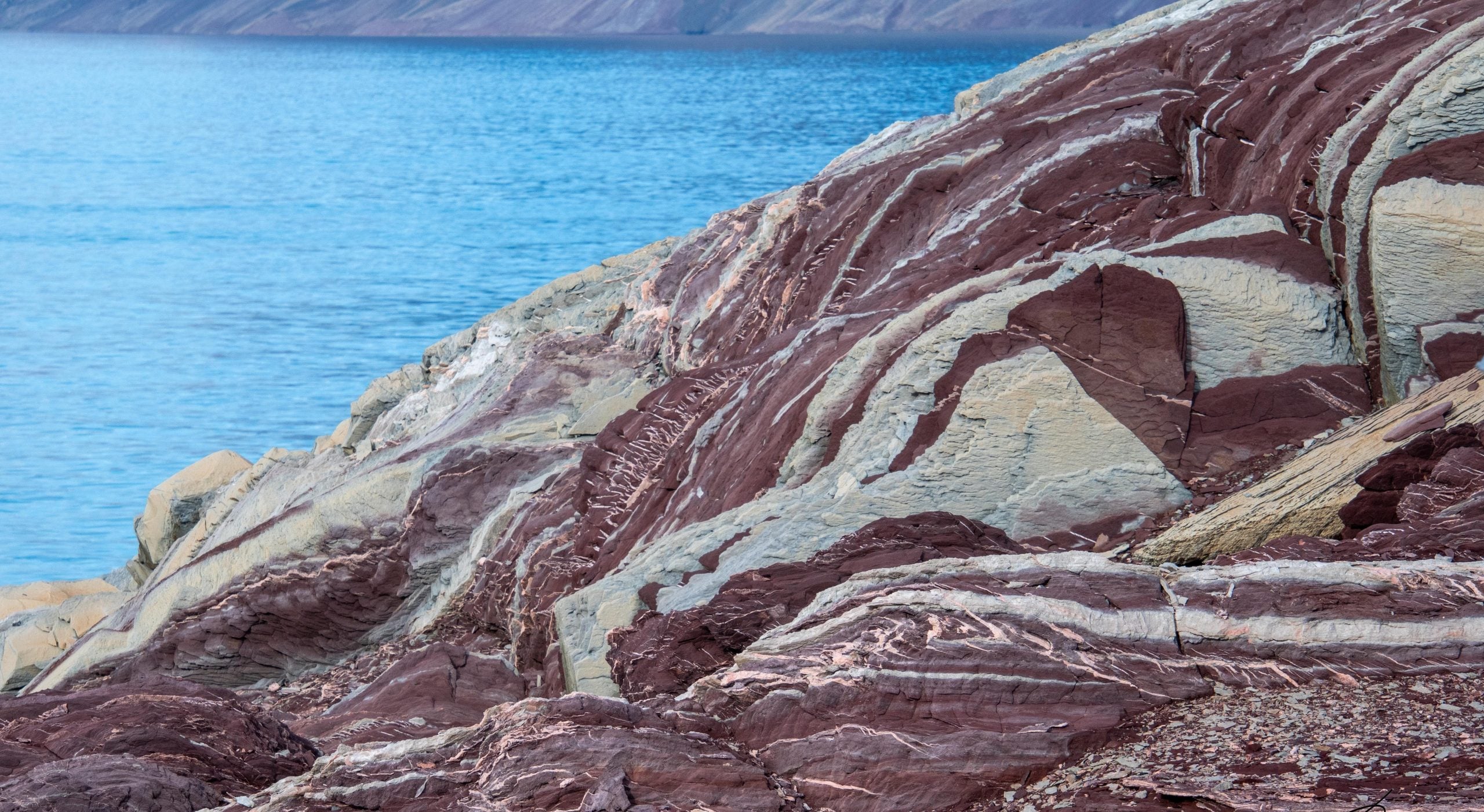 Layered red and beige sedimentary rock formations along a blue body of water.