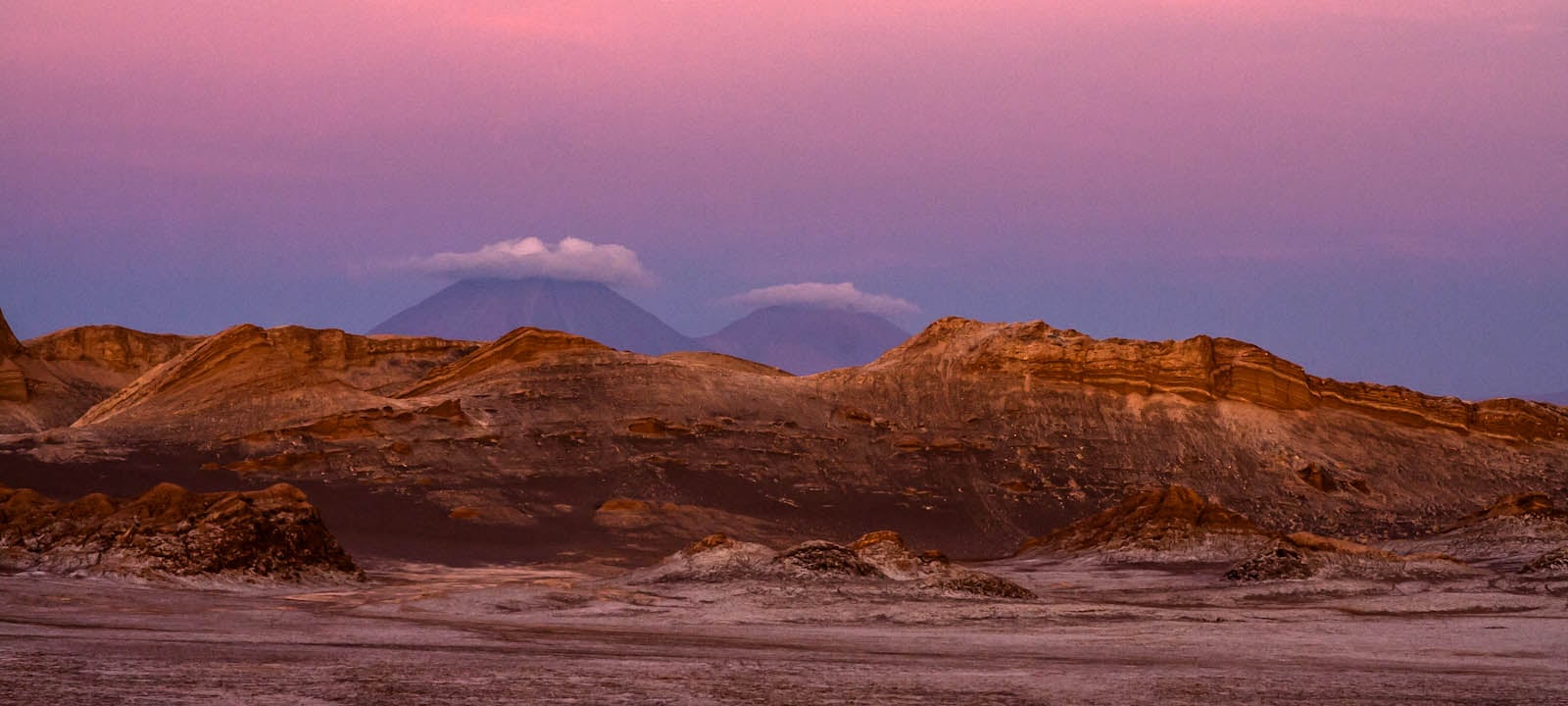 Rugged desert landscape with rocky ridges under a purple twilight sky and two distant mountain peaks capped with clouds.