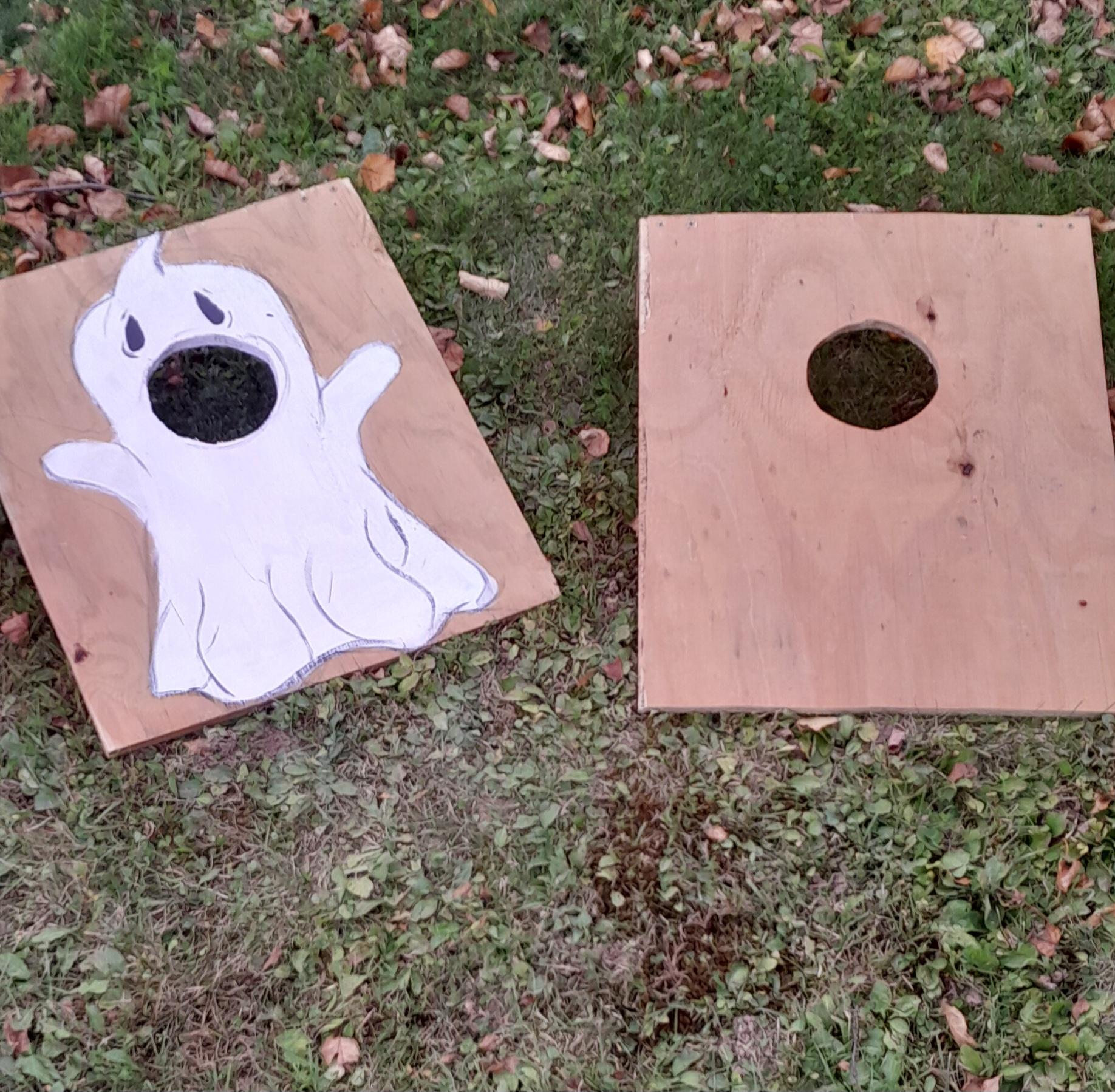 Two wooden cornhole boards resting on grass, one featuring a painted white ghost with a cutout hole as its mouth.