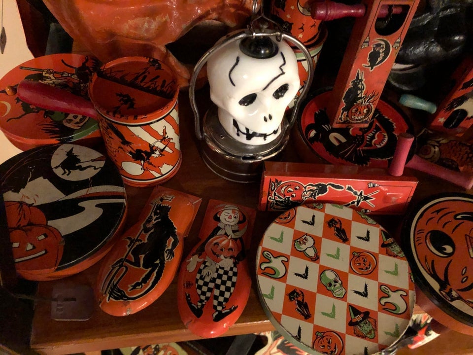 A collection of vintage Halloween-themed tin toys and containers, including a white skull lantern, a checkered plate with pumpkin designs, and various metal figures featuring witches and black cats.