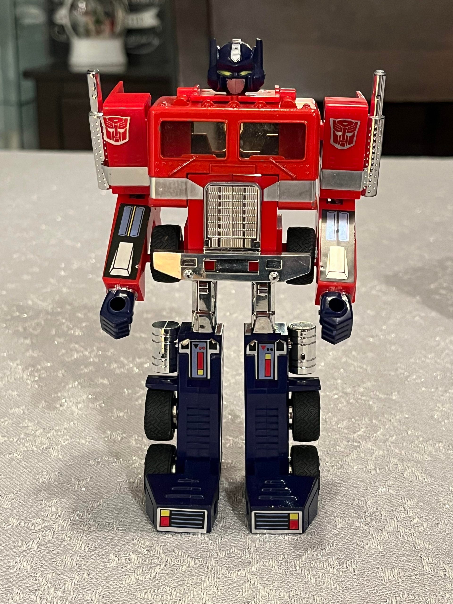 A red and blue robot toy, resembling a classic Optimus Prime Transformer, stands upright on a textured gray surface with details like chrome accents and decals visible.