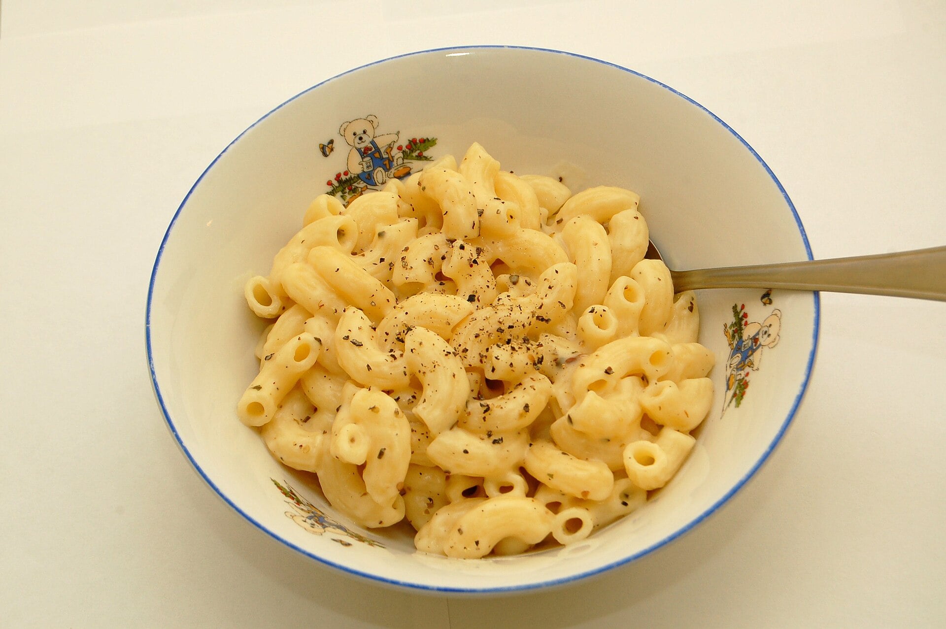 A bowl of macaroni and cheese topped with black pepper, served in a white bowl with bear illustrations, with a spoon inside.