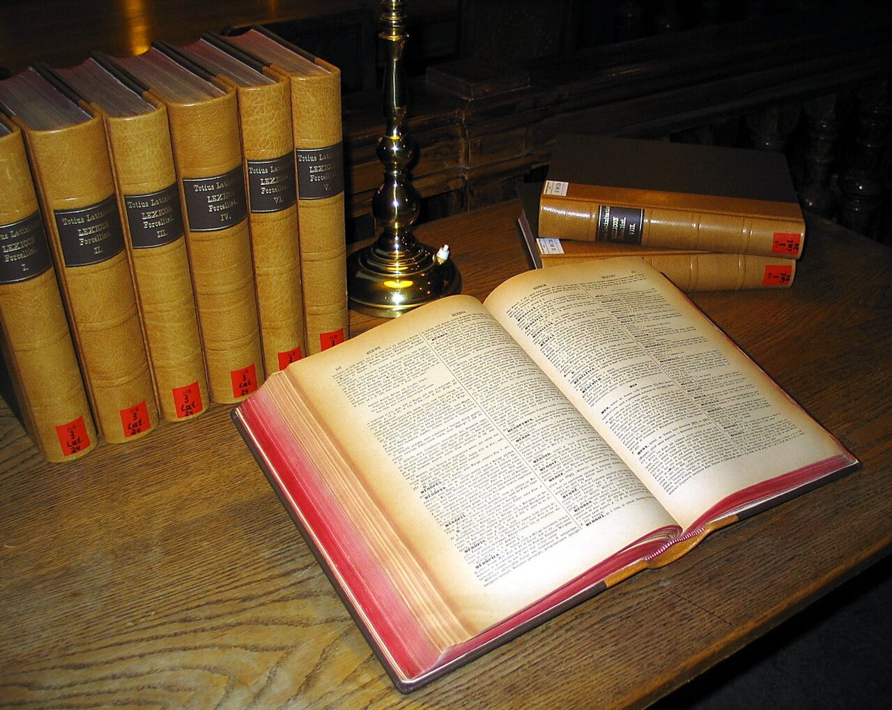 An open reference book lies on a wooden desk next to several leather-bound volumes and a brass lamp.
