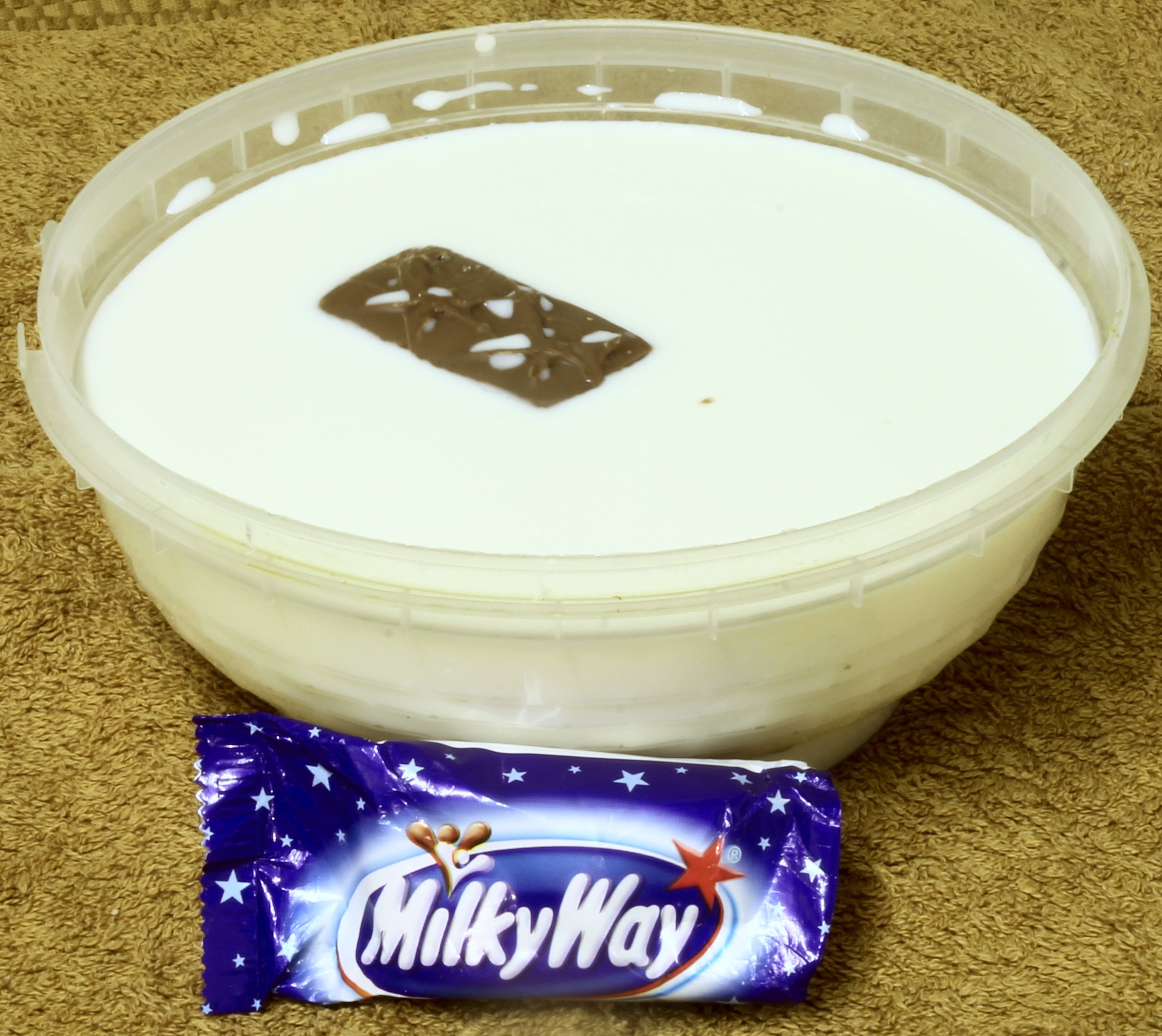 A plastic bowl filled with white liquid featuring a chocolate bar fragment on the surface, with a sealed Milky Way candy bar placed in front.