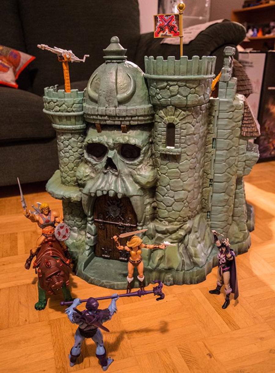 Four action figures with weapons stand in front of a green, castle-shaped toy with a skull entrance and a flag on top, set on a wooden floor in a living room.