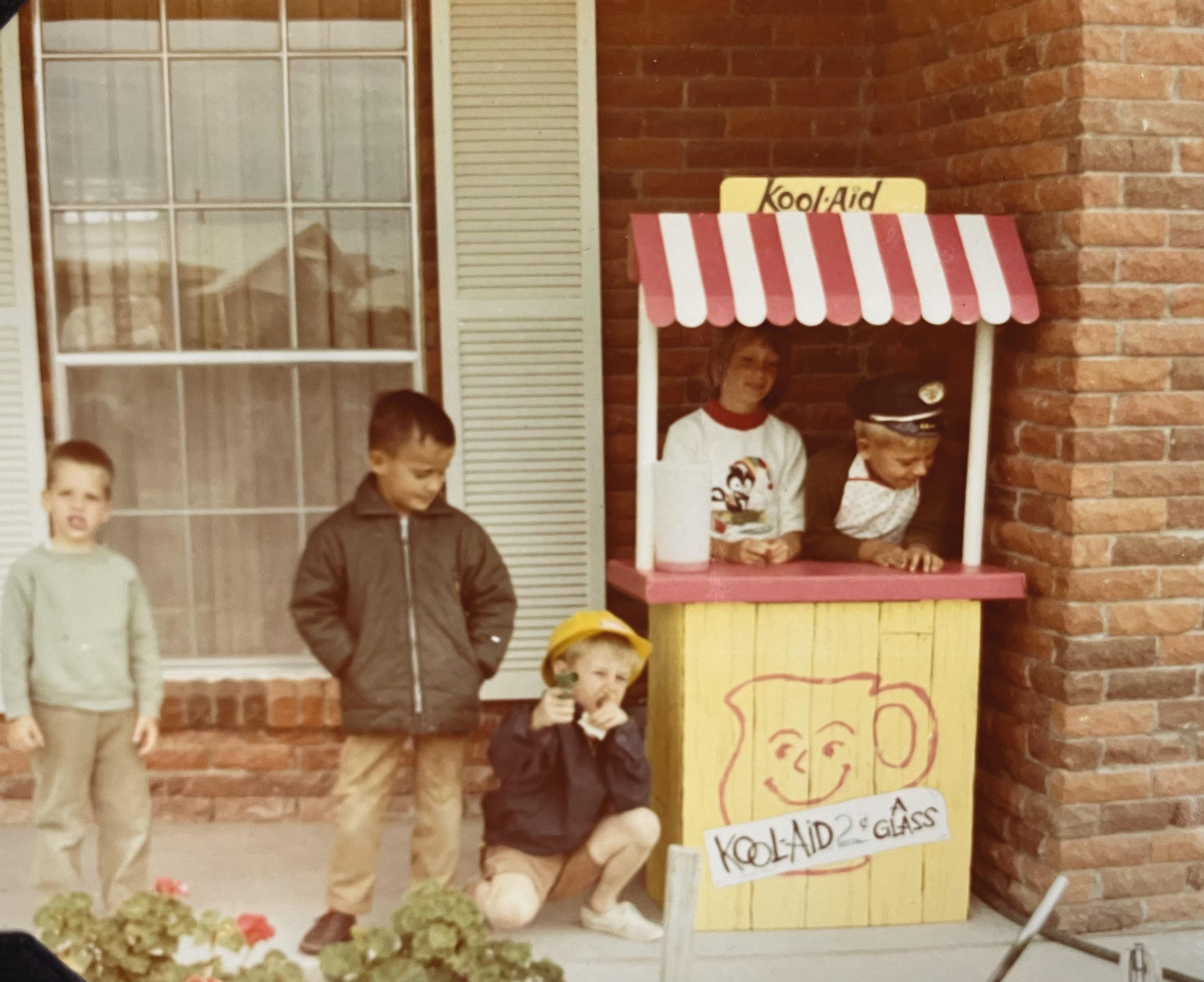 Four children playing at a homemade Kool-Aid stand in front of a brick house.
