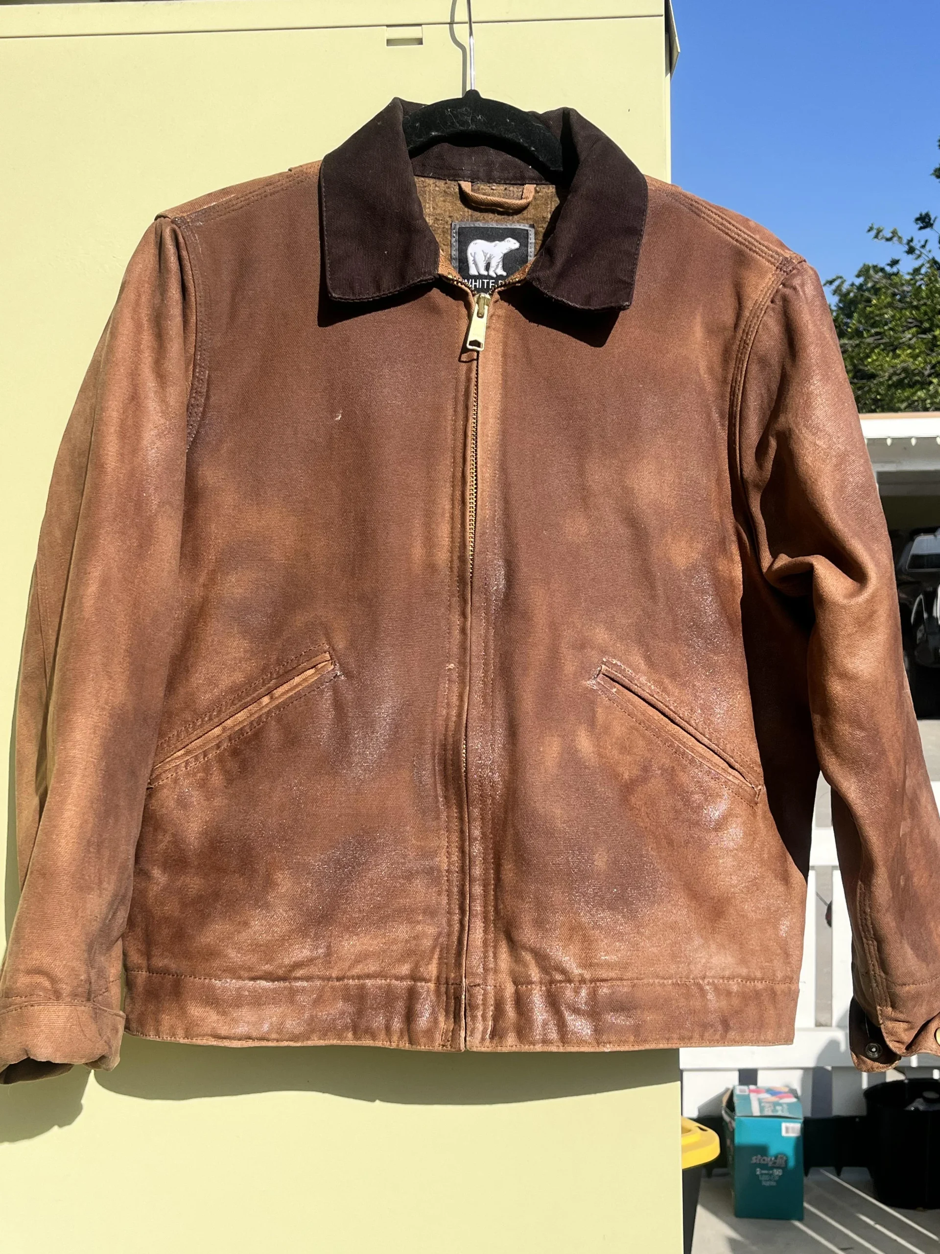 A brown leather jacket with a dark collar hangs on a hanger outside against a light green wall, with sunlight highlighting its worn texture.